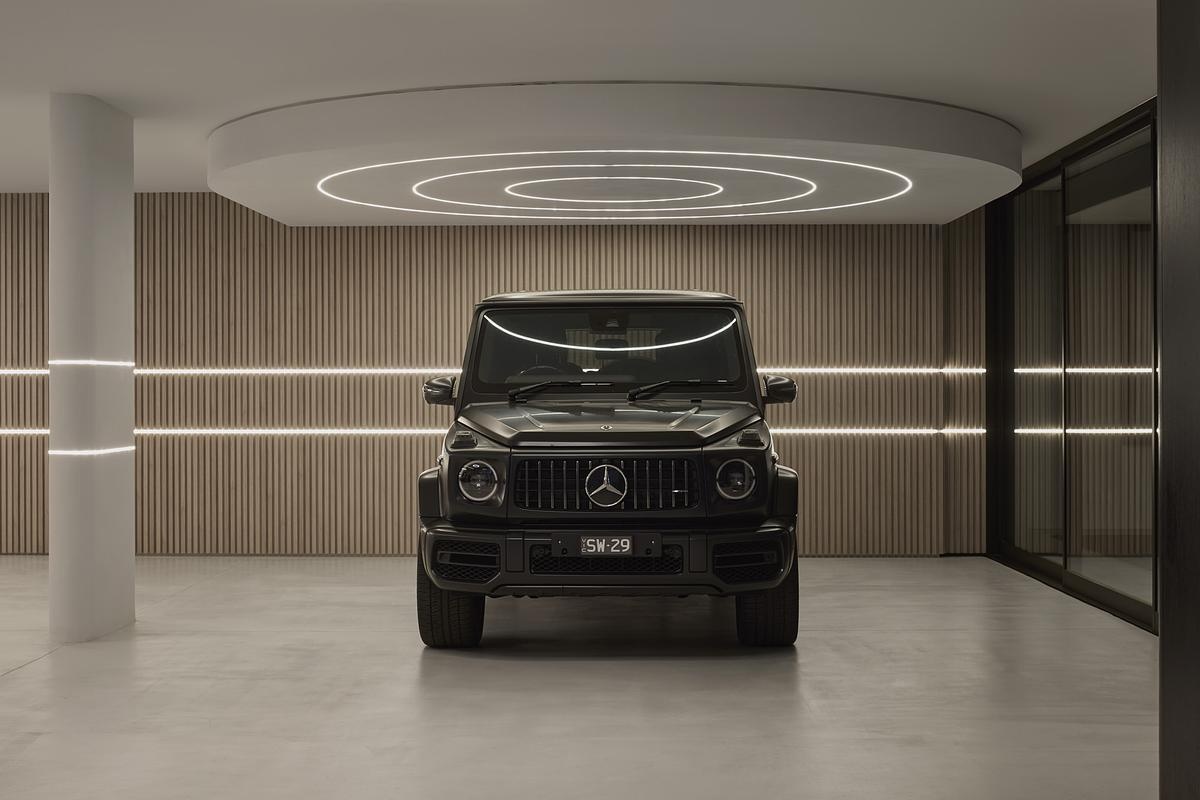The garage feels more like a luxury showroom than a parking spot. Soft wood paneling lines the walls in gentle vertical slats, warmed by sleek bands of LED lighting that wrap the space in a futuristic glow. Circular light patterns on the ceiling draw attention to the centerpiece below, while polished concrete floors keep the mood clean and grounded.