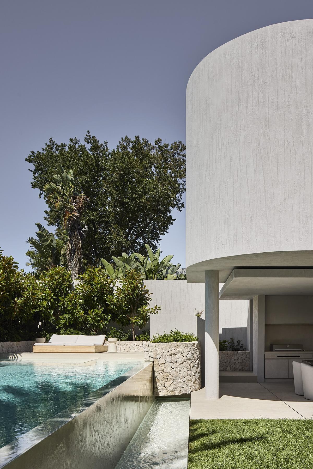 A micro-cement infinity pool glimmers just beyond the patio, visually extending the home’s serene interiors into the lush garden designed by Jack Merlo. Patterned paving and feature rock cladding frame the pool area, where indoor-outdoor living is experienced at its most luxurious.
