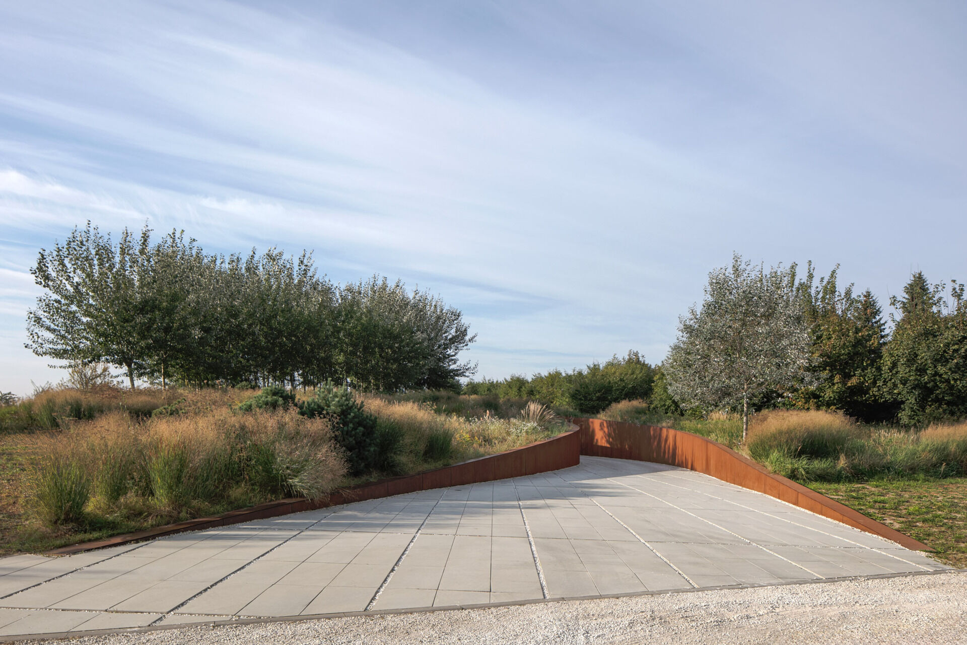 The entrance to the home is cut into the slope like a subtle steel-lined ravine. This sunken driveway leads to a small, recessed patio and front door, designed to keep the view concealed until entry. The use of corten steel along the driveway and facade gives the entrance a grounded, weathered character that blends with the terrain.