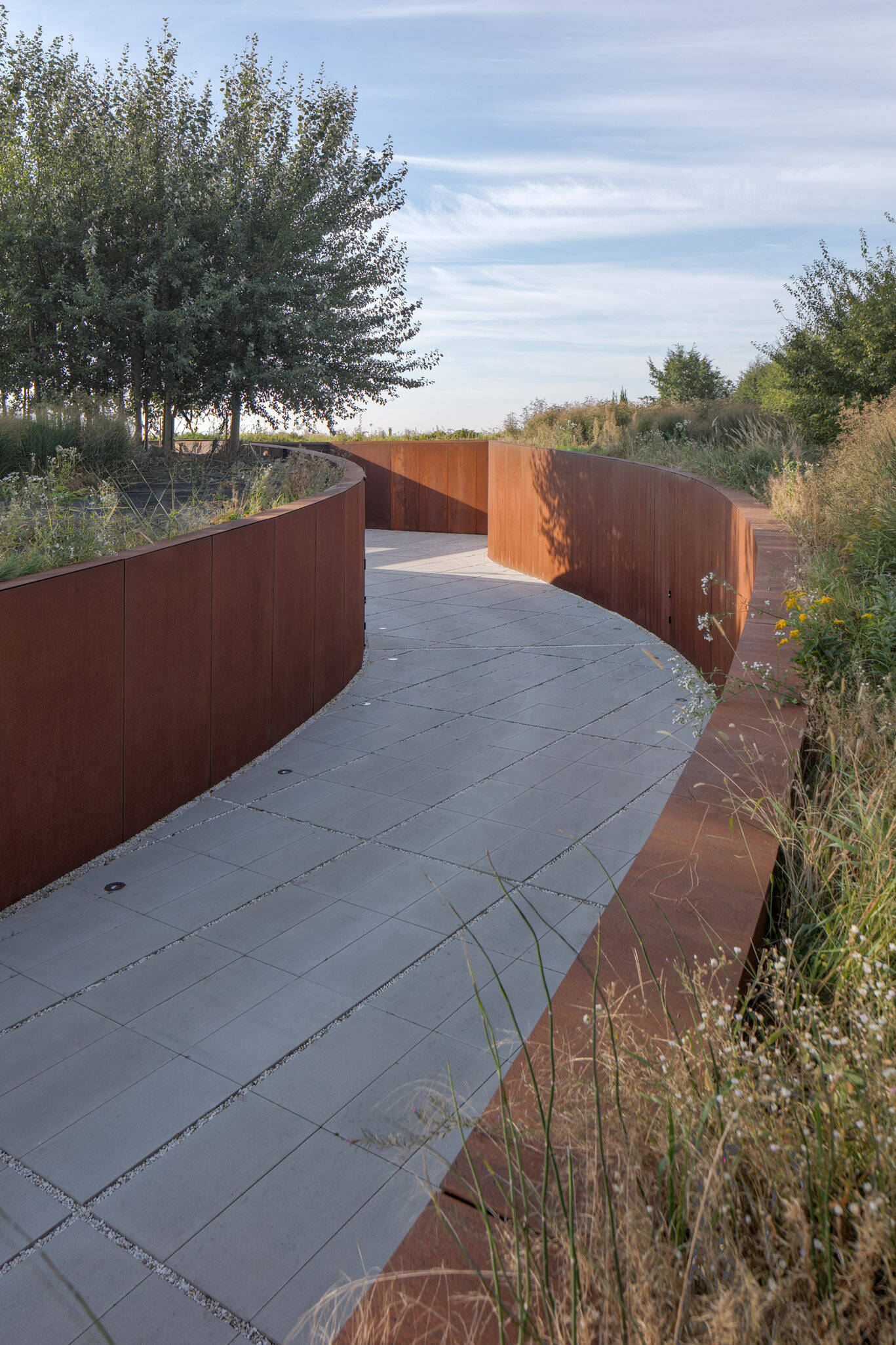 The entrance to the home is cut into the slope like a subtle steel-lined ravine. This sunken driveway leads to a small, recessed patio and front door, designed to keep the view concealed until entry. The use of corten steel along the driveway and facade gives the entrance a grounded, weathered character that blends with the terrain.