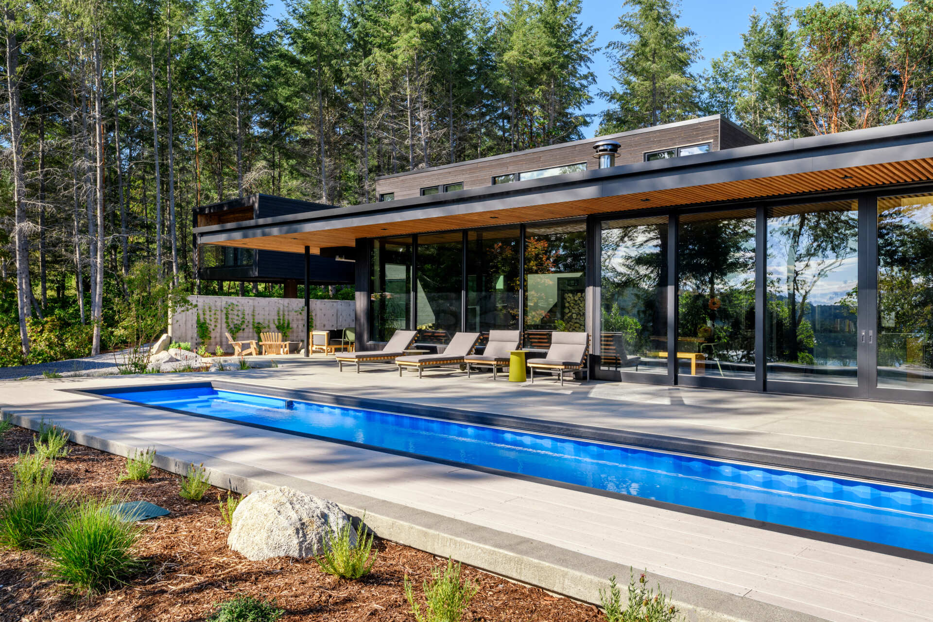 A forest-framed patio with a narrow lap pool, slatted ceiling, firepit, and warm wood details, designed for quiet mornings and slow evenings outdoors.