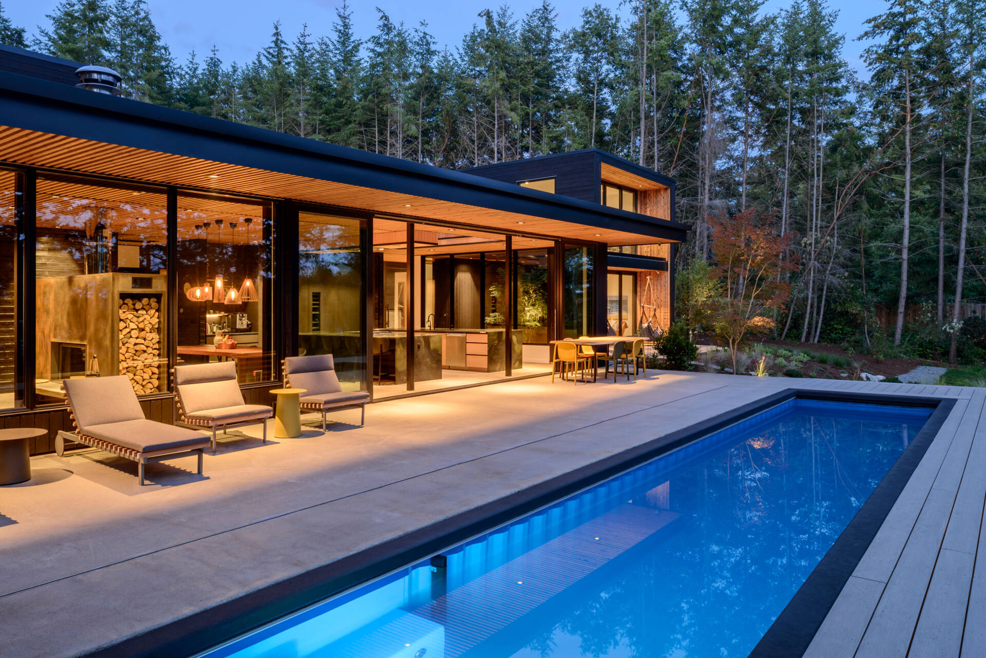 A forest-framed patio with a narrow lap pool, slatted ceiling, firepit, and warm wood details, designed for quiet mornings and slow evenings outdoors.
