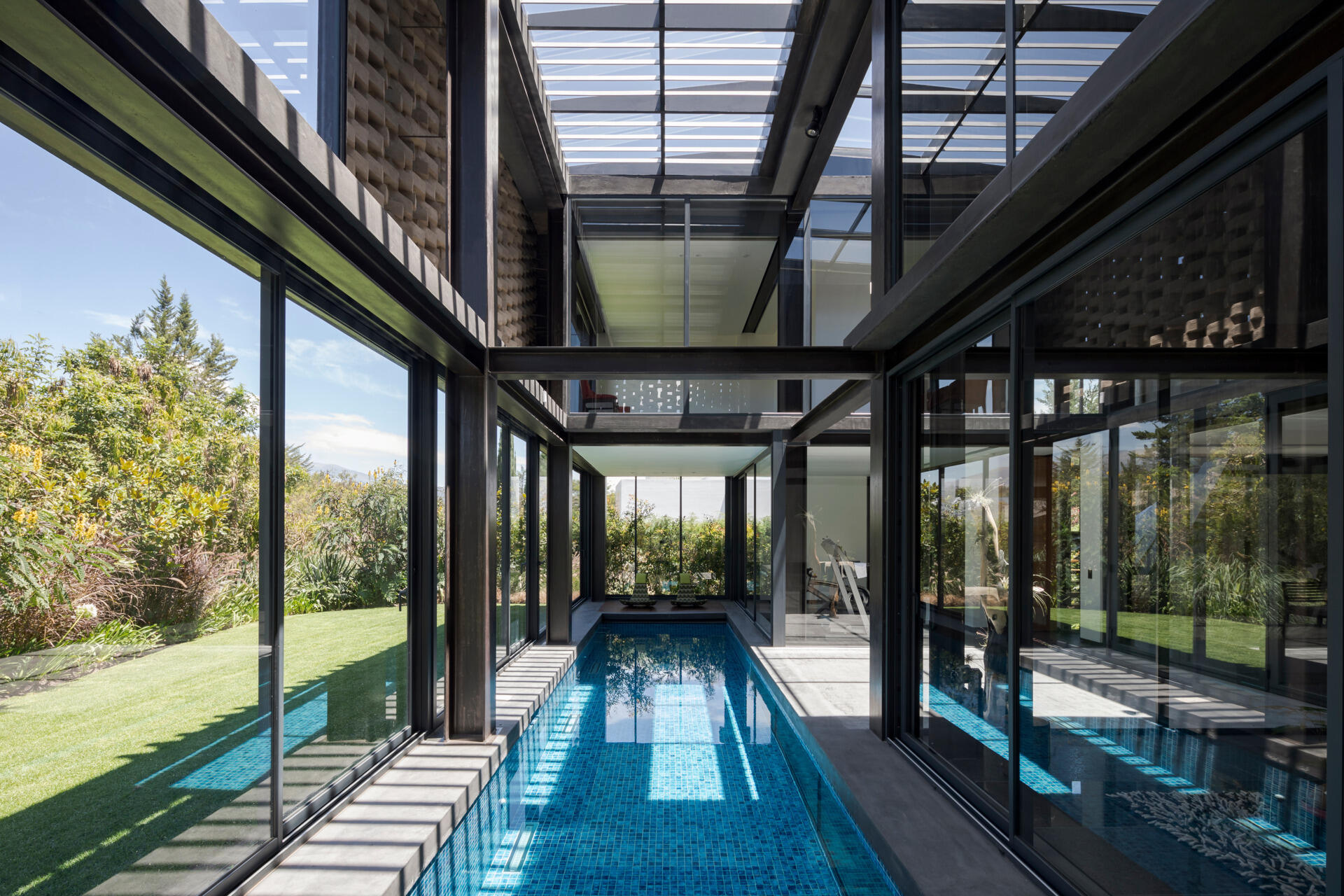At the upper level, a sleek pool stretches beside a flat lawn. Floor-to-ceiling glass dissolves the boundary between inside and out, uniting water, architecture, and landscape.