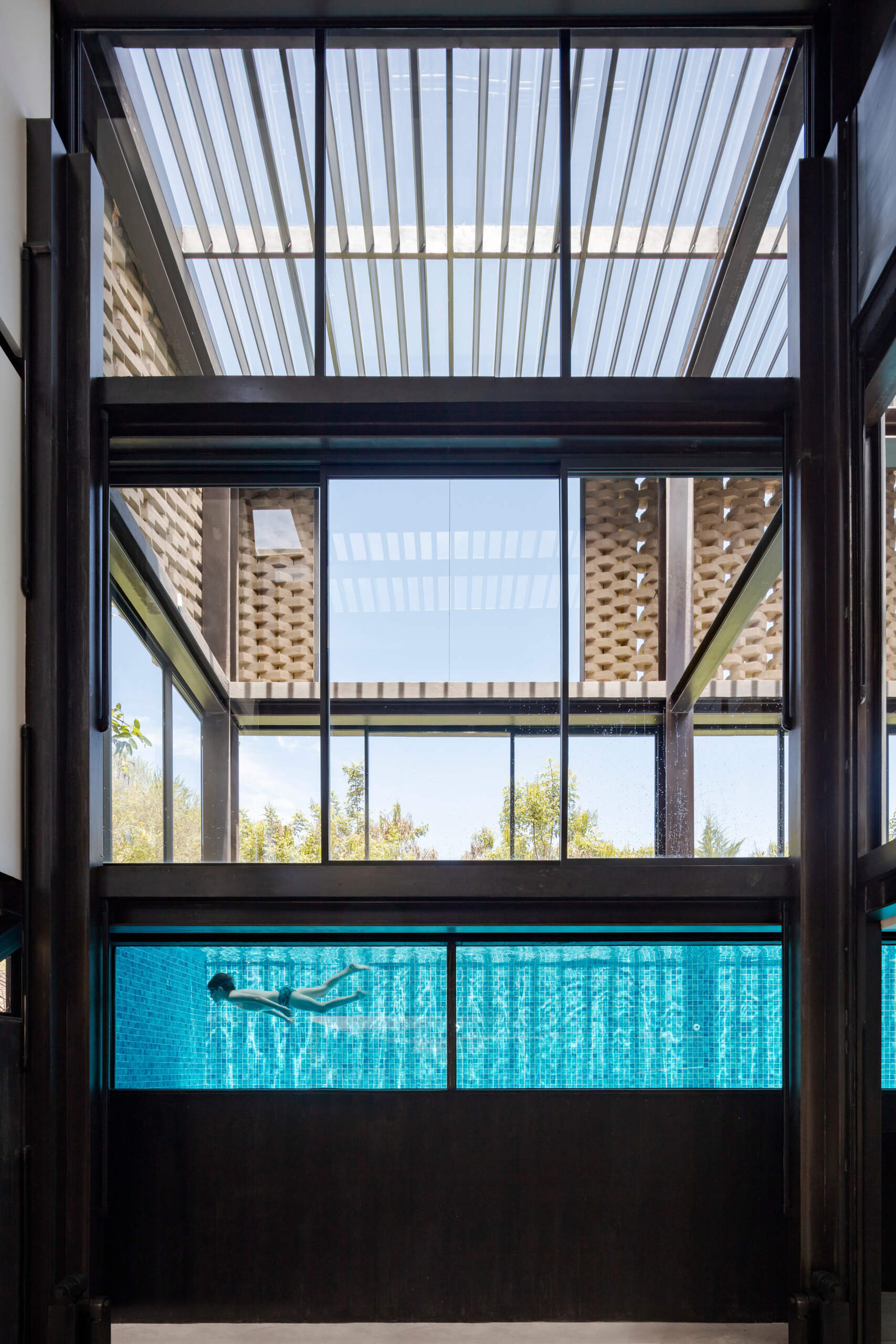 At the upper level, a sleek pool stretches beside a flat lawn. Floor-to-ceiling glass dissolves the boundary between inside and out, uniting water, architecture, and landscape.