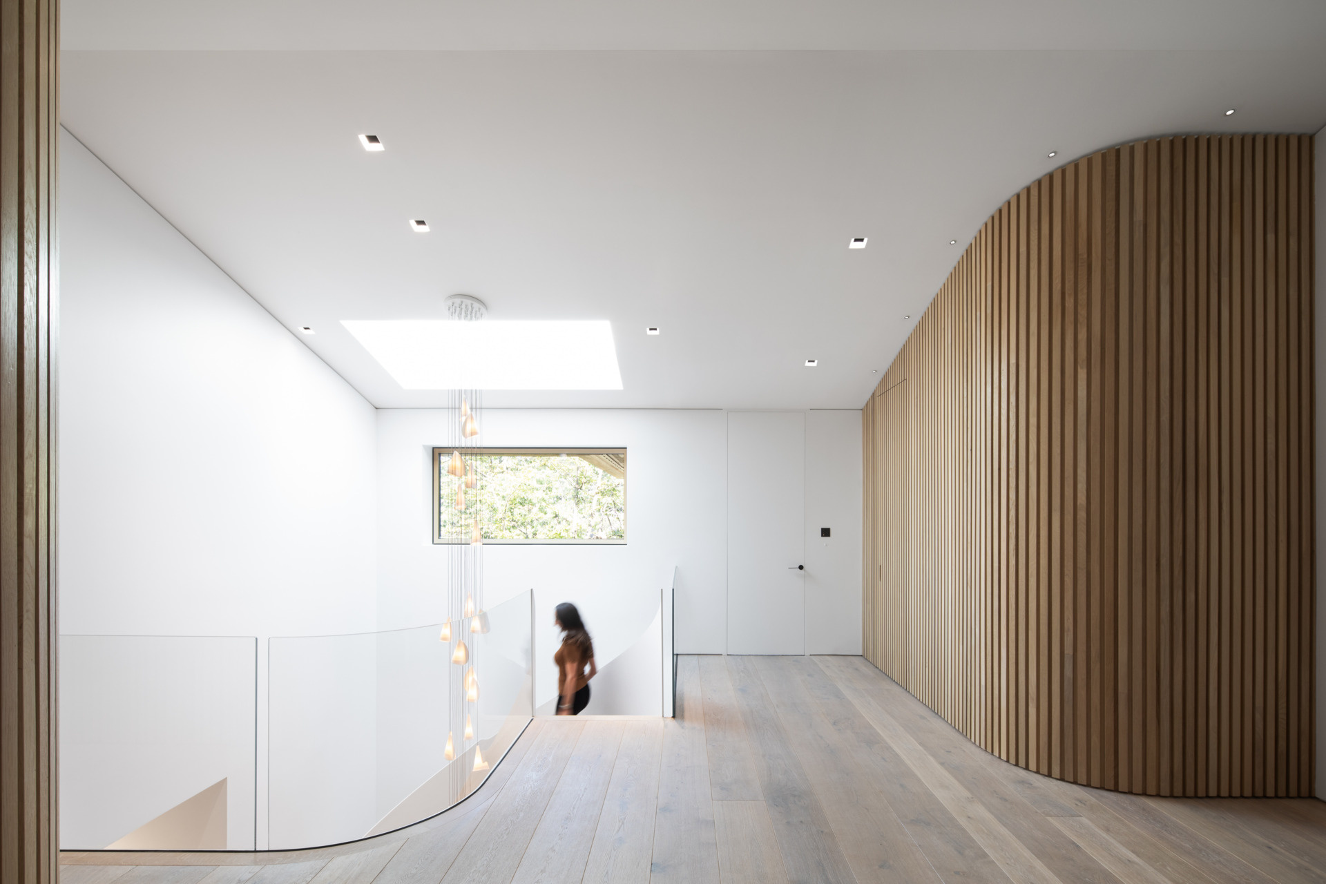 Light-filled and open, the landing at the top of the stairs offers a graceful transition between levels. A curved slatted wall flows into pale wood flooring, creating a gentle sense of movement. Natural light pours in through a skylight and side window, illuminating the soft pendant lights that cascade down the stairwell.