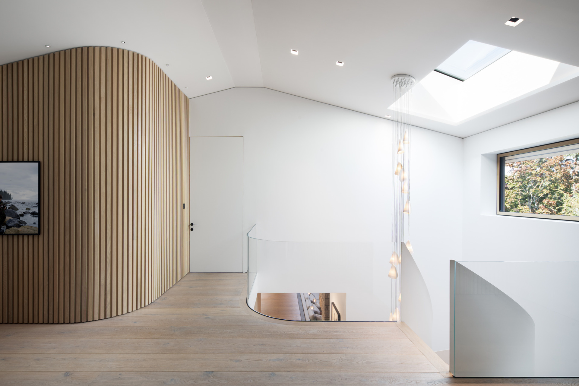 Light-filled and open, the landing at the top of the stairs offers a graceful transition between levels. A curved slatted wall flows into pale wood flooring, creating a gentle sense of movement. Natural light pours in through a skylight and side window, illuminating the soft pendant lights that cascade down the stairwell.