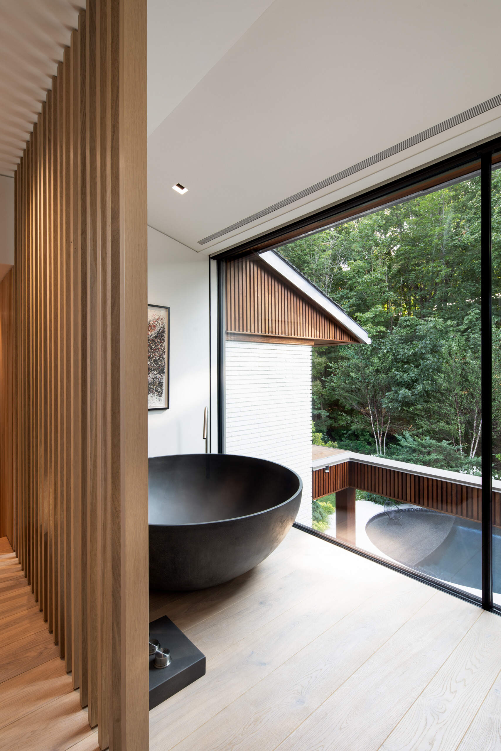 A unexpected area offers a moment of quiet luxury, where a sculptural black tub anchors the space. Framed by pale wood slats and floor-to-ceiling glass, it balances privacy with openness. Treetops sway just beyond the glass, and soft light filters in, creating the feeling of a serene retreat.