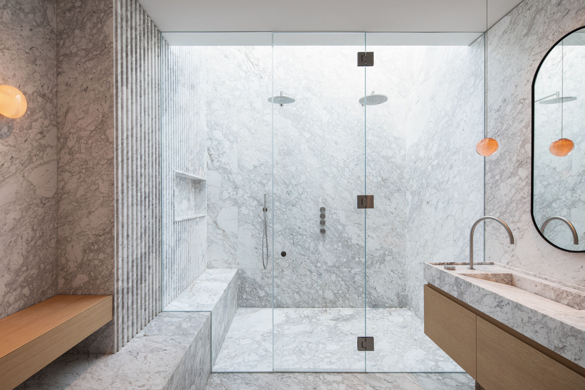 A luxurious bathroom is enveloped in cloud-like marble, creating a calm, spa-like retreat. A spacious walk-in shower with frameless glass lets natural light shine through, while subtle vertical grooves in the stone add gentle texture. Warm wood elements in the bench and vanity offer a natural contrast, while delicate pendant lights and a curved mirror soften the space, making it feel peaceful and restful.