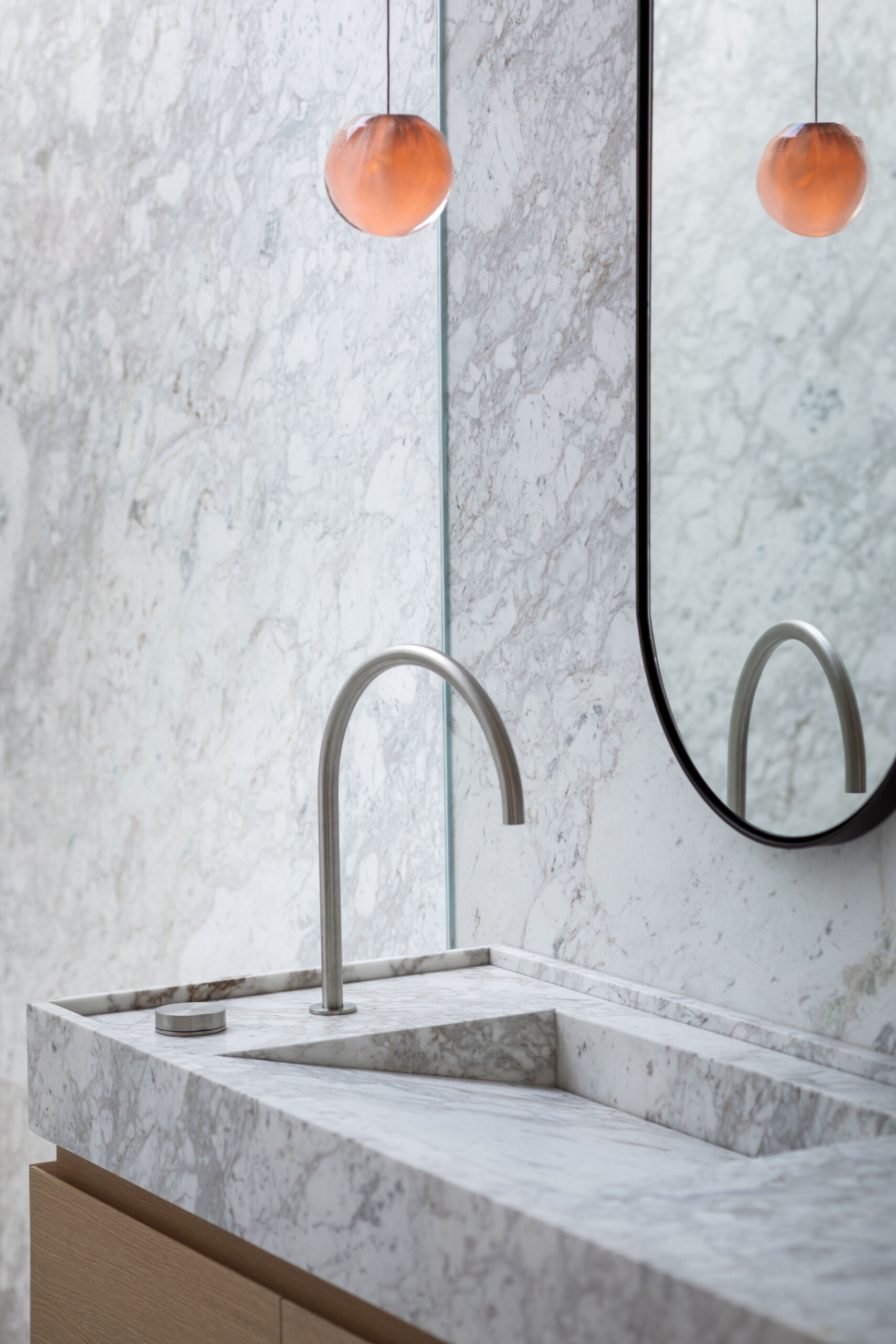 A luxurious bathroom is enveloped in cloud-like marble, creating a calm, spa-like retreat. A spacious walk-in shower with frameless glass lets natural light shine through, while subtle vertical grooves in the stone add gentle texture. Warm wood elements in the bench and vanity offer a natural contrast, while delicate pendant lights and a curved mirror soften the space, making it feel peaceful and restful.