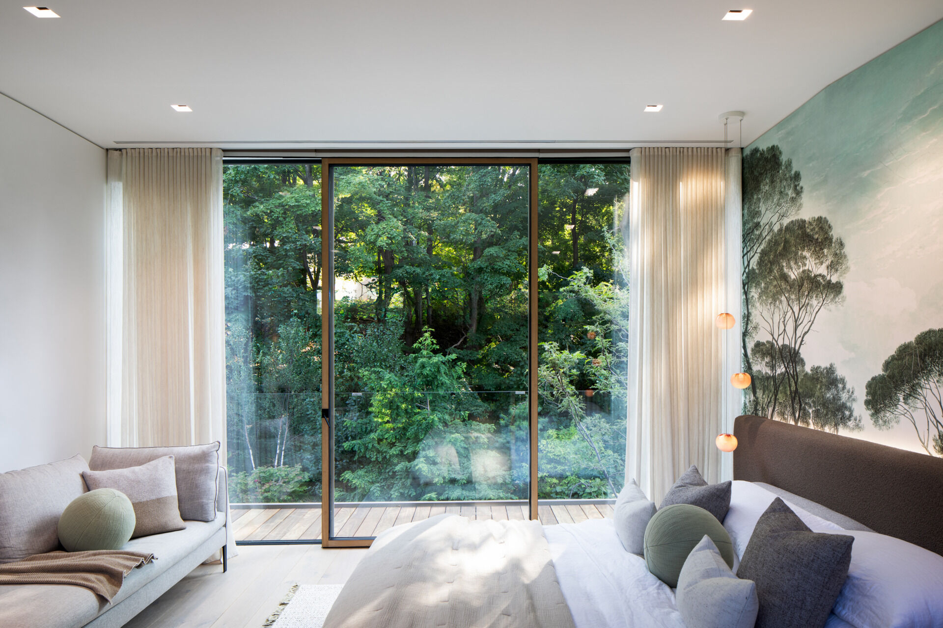 With sheer curtains and floor-to-ceiling glass, this bedroom opens to a lush tree canopy, offering a serene connection to nature. Soft, layered textiles in muted earth tones reflect the calming colors of the mural behind the bed, where delicate tree silhouettes add a gentle, nature-inspired accent.