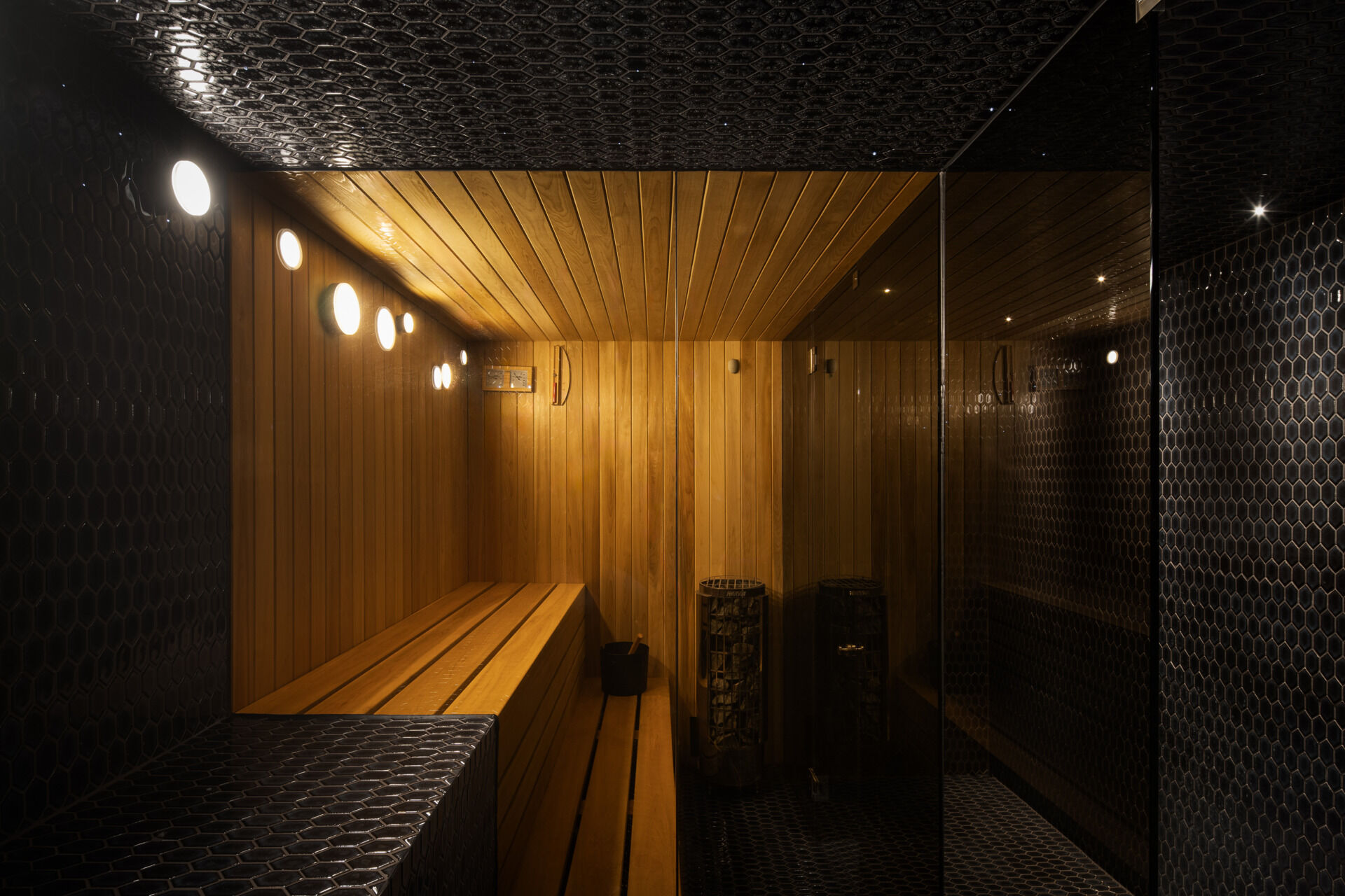 A sauna balances warmth and contrast, where vertical wood paneling meets glossy black tile for a modern, grounded feel. Soft circular lighting adds a gentle rhythm, highlighting the clean lines and natural materials.