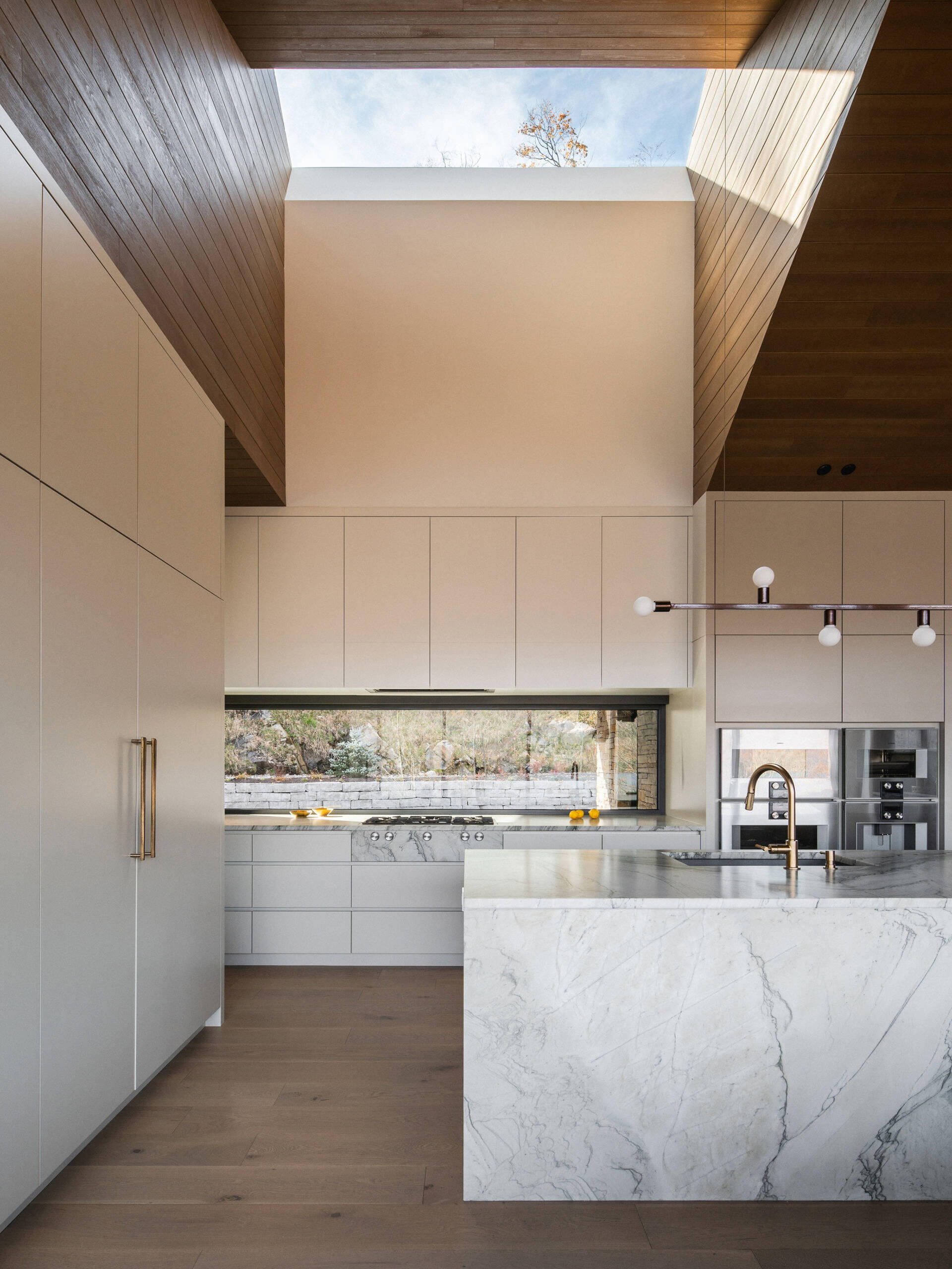 The ceiling rises over the kitchen to a skylight, increasing light and volume. A large island anchors the space, while a horizontal window above the counter provides a framed view of the forest. Behind a paneled wall, a separate pantry kitchen adds functionality without disrupting the open layout.