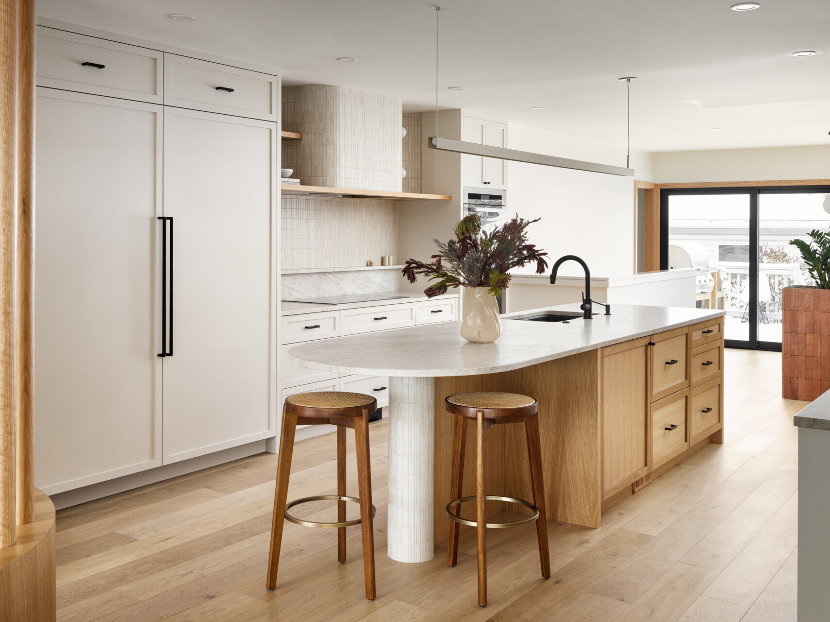 At the heart of this home lies the redesigned kitchen with neutral cabinetry and countertops to reflect light and stretch sightlines. A warm wood island blends into the hardwood flooring, encouraging a natural visual flow toward the rear windows. A curved hood fan and island counter softens the typical edges of kitchen details and continuing the home’s curvilinear language.