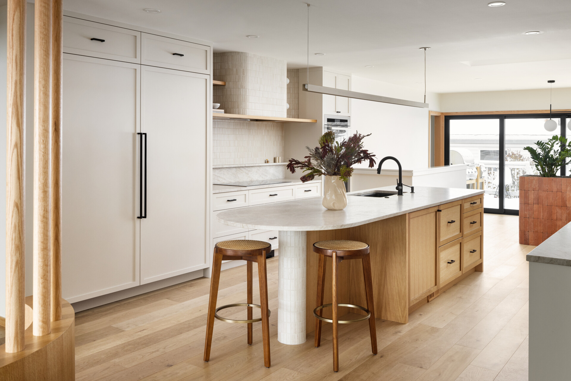 At the heart of this home lies the redesigned kitchen with neutral cabinetry and countertops to reflect light and stretch sightlines. A warm wood island blends into the hardwood flooring, encouraging a natural visual flow toward the rear windows. A curved hood fan and island counter softens the typical edges of kitchen details and continuing the home’s curvilinear language.