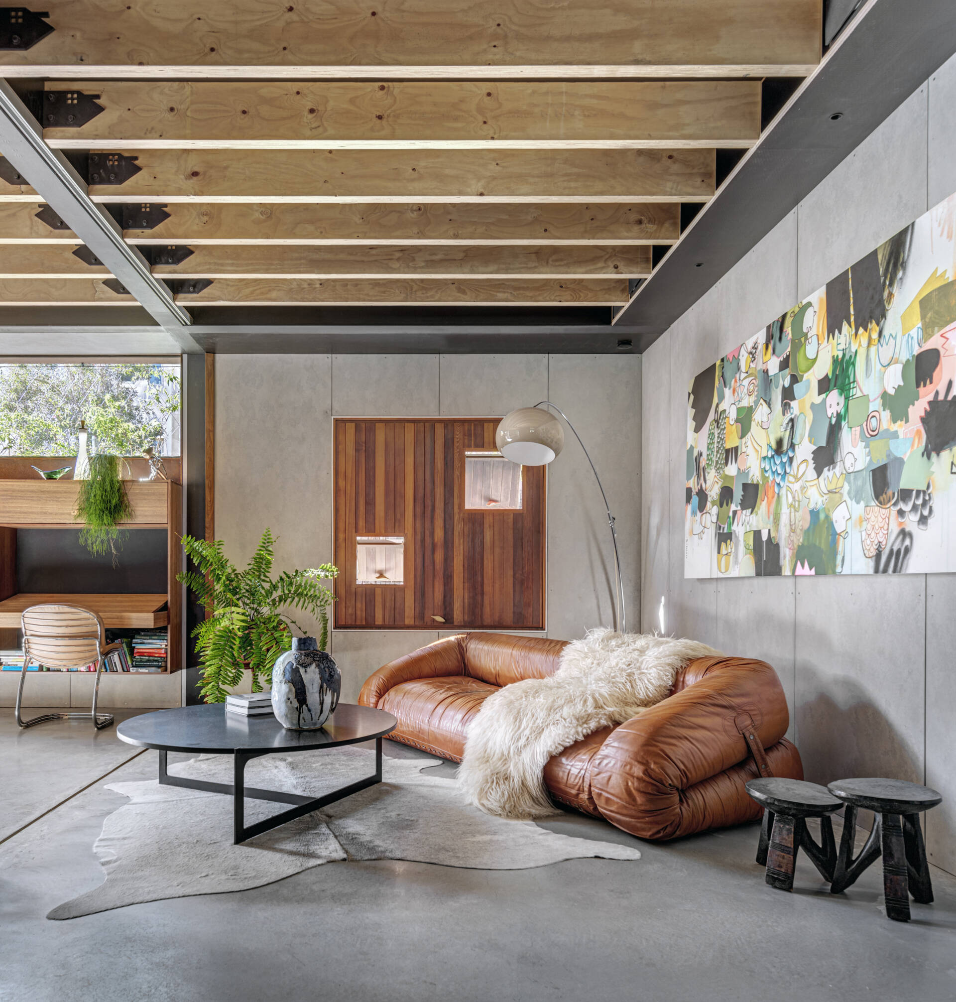 A living room balances modern simplicity with inviting warmth. A leather sofa anchors the space, while an abstract painting adds subtle color against concrete walls. Soft natural textures, like a sheepskin throw and leafy fern, temper the industrial feel of exposed beams. Light filters through a timber opening, casting a warm glow that elevates everyday comfort.
