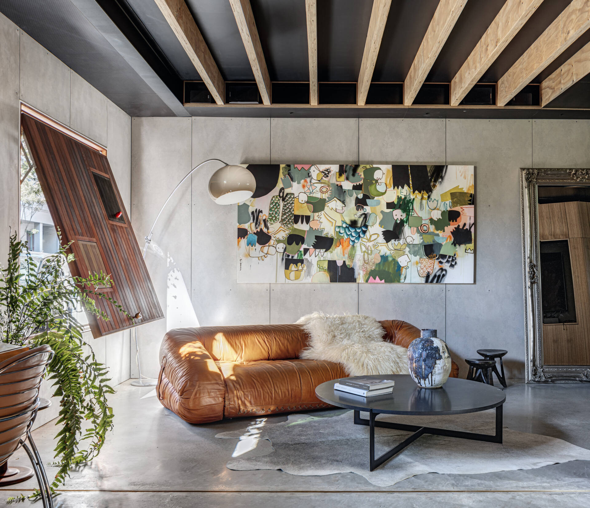 A living room balances modern simplicity with inviting warmth. A leather sofa anchors the space, while an abstract painting adds subtle color against concrete walls. Soft natural textures, like a sheepskin throw and leafy fern, temper the industrial feel of exposed beams. Light filters through a timber opening, casting a warm glow that elevates everyday comfort.