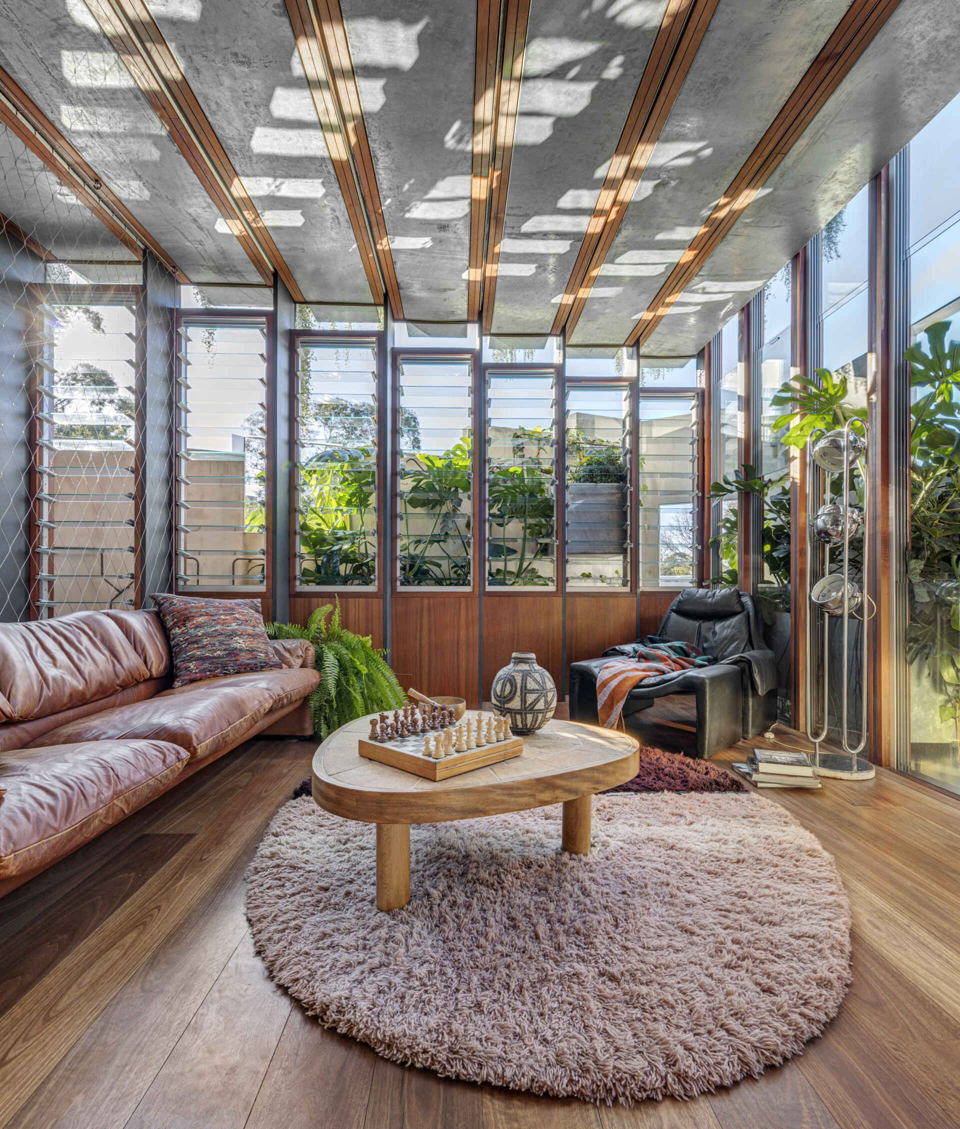 Bathed in dappled sunlight, this secondary living room / sunroom feels like a peaceful retreat with its soft leather sofa and plush circular rug underfoot. The natural wood coffee table is ready for a quiet cup of tea, while leafy plants thrive by the tall louvered windows. Warm tones and rich textures balance the crisp, modern lines of the concrete ceiling, creating a space that feels perfect for unwinding and soaking in a slow afternoon.