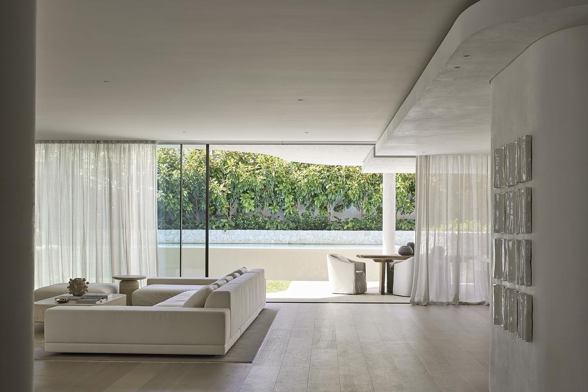 Soft natural light pours through sheer curtains into this serene, open-concept living space. The neutral palette of creamy whites and pale wood flooring creates a calming flow from the lounge to the dining nook, while curved architectural lines add a sculptural touch. Floor-to-ceiling glass blurs the boundary between indoors and out, offering a peaceful view of lush greenery and pool.