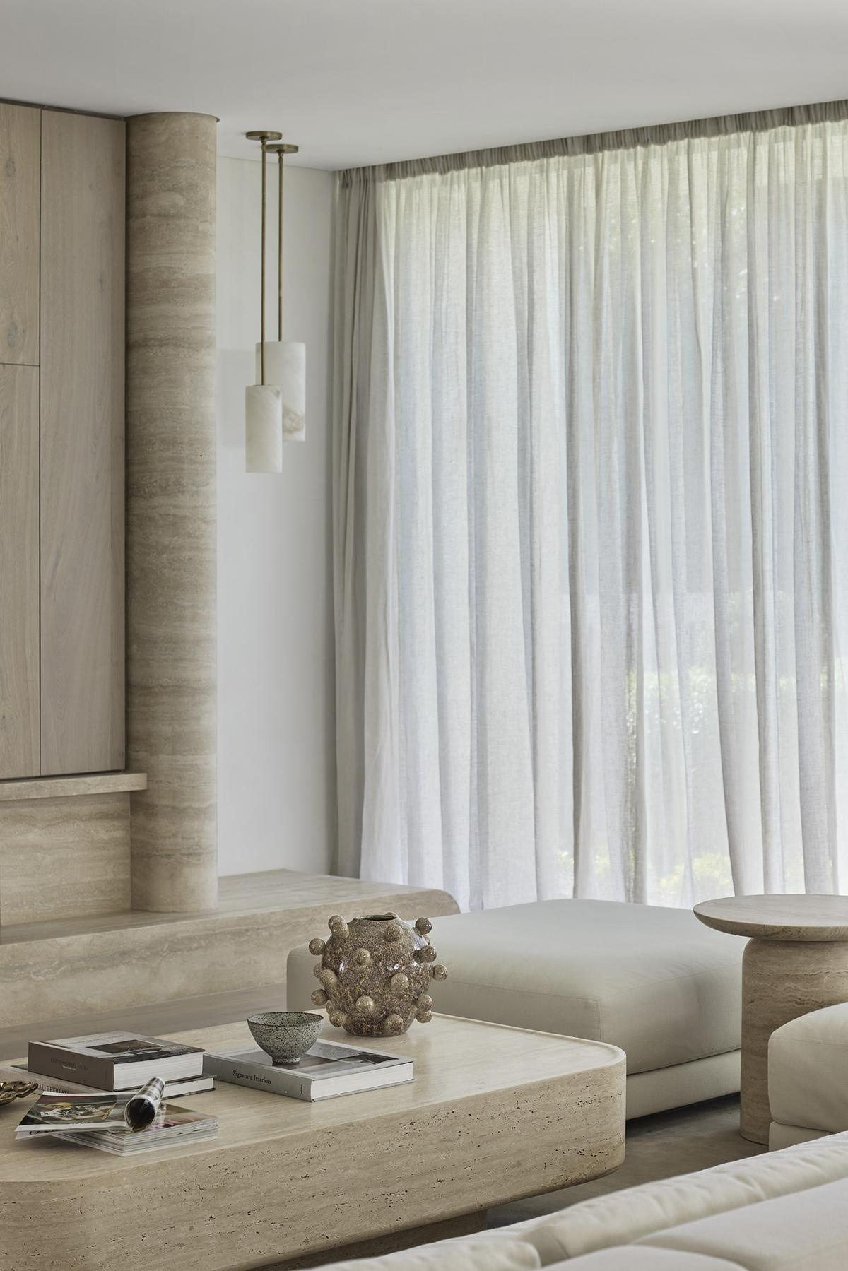 Soft natural light pours through sheer curtains into this serene, open-concept living space. The neutral palette of creamy whites and pale wood flooring creates a calming flow from the lounge to the dining nook, while curved architectural lines add a sculptural touch. Floor-to-ceiling glass blurs the boundary between indoors and out, offering a peaceful view of lush greenery and pool.