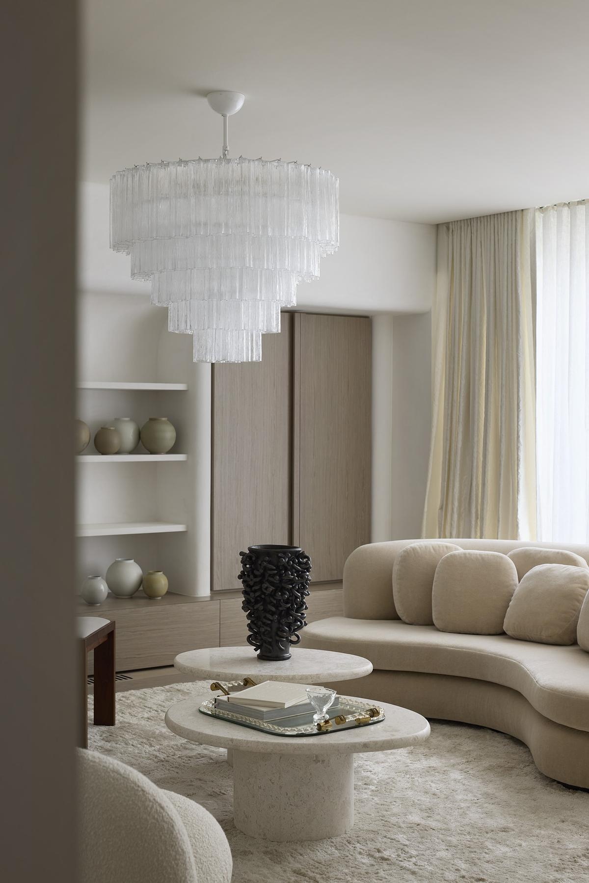 Plush textures and sculptural forms shape this softly lit lounge, where curved seating invites relaxed conversation. A layered chandelier catches light through creamy drapes, while stone tables and a shag rug add warmth. Nearby, a sleek black fireplace with nearby wood storage brings rustic charm.