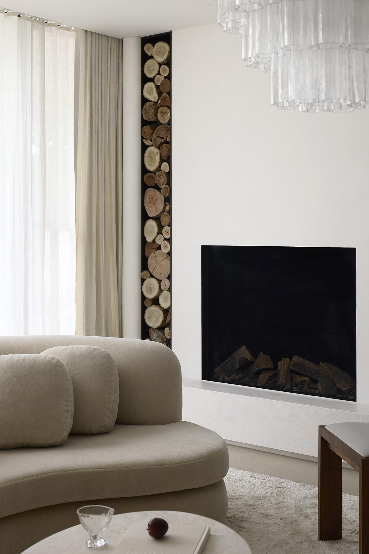 Plush textures and sculptural forms shape this softly lit lounge, where curved seating invites relaxed conversation. A layered chandelier catches light through creamy drapes, while stone tables and a shag rug add warmth. Nearby, a sleek black fireplace with nearby wood storage brings rustic charm.