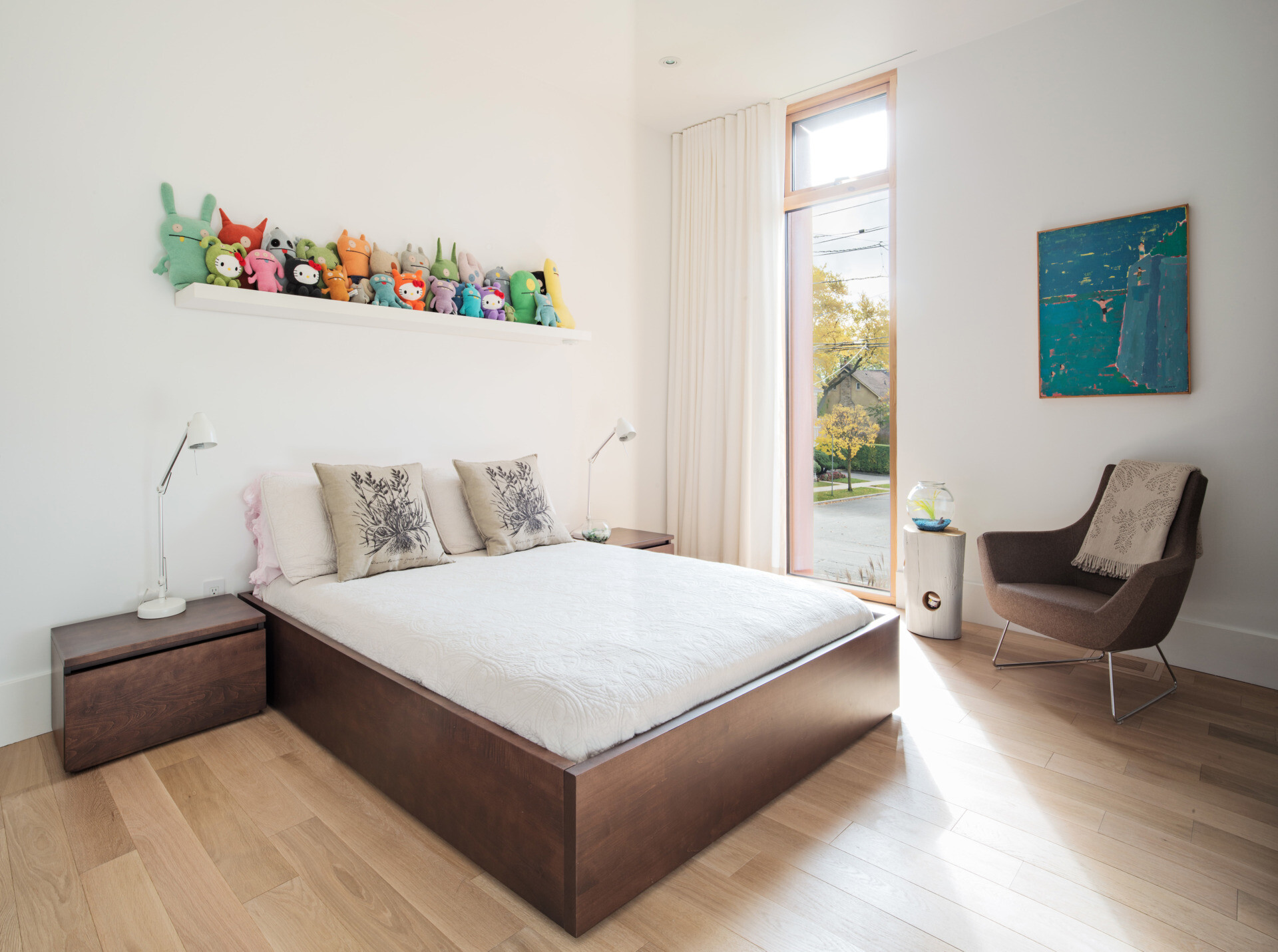 A children’s bedroom is bright and airy, with natural light streaming in through the window. Pops of color come from playful artwork and a collection of plush toys neatly arranged on the shelf.