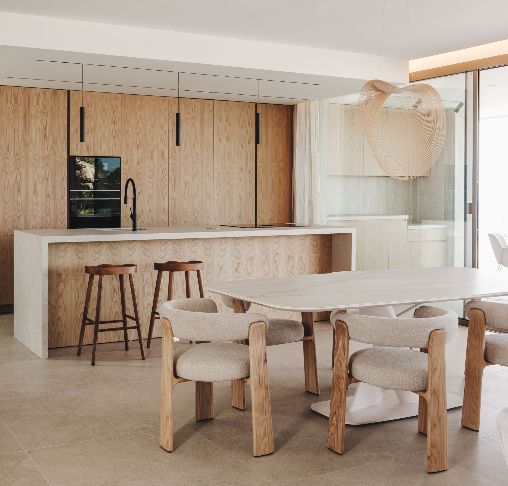 In this open plan dining area, a pale oak table and sculptural chairs sit beneath a soft pendant, framing views and continuing the home's natural palette.