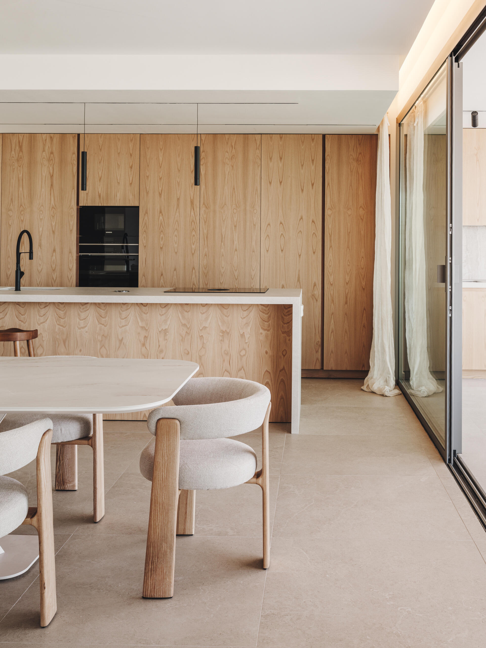 In this open plan dining area, a pale oak table and sculptural chairs sit beneath a soft pendant, framing views and continuing the home's natural palette.