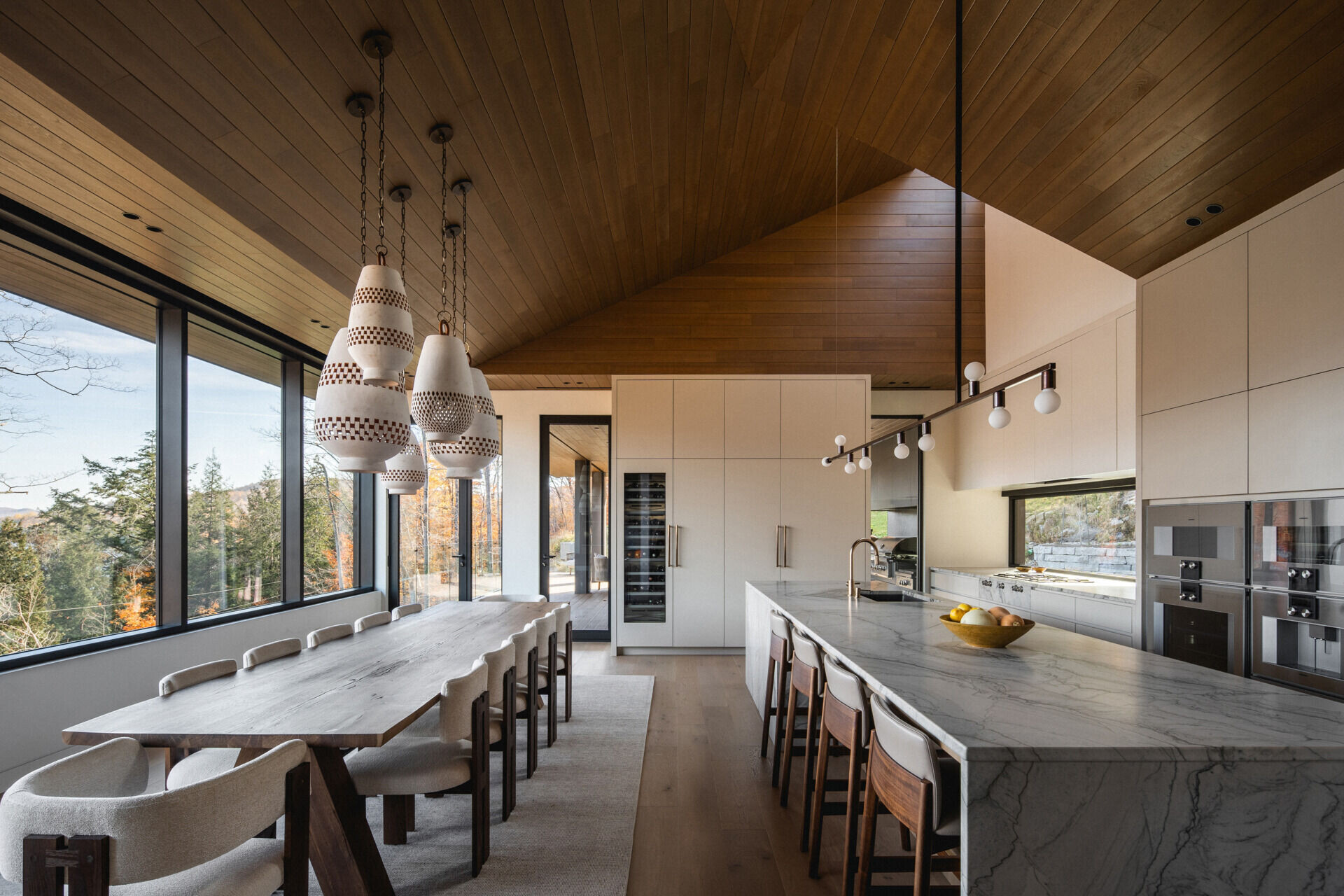 The ceiling rises over the kitchen to a skylight, increasing light and volume. A large island anchors the space, while a horizontal window above the counter provides a framed view of the forest. Behind a paneled wall, a separate pantry kitchen adds functionality without disrupting the open layout.