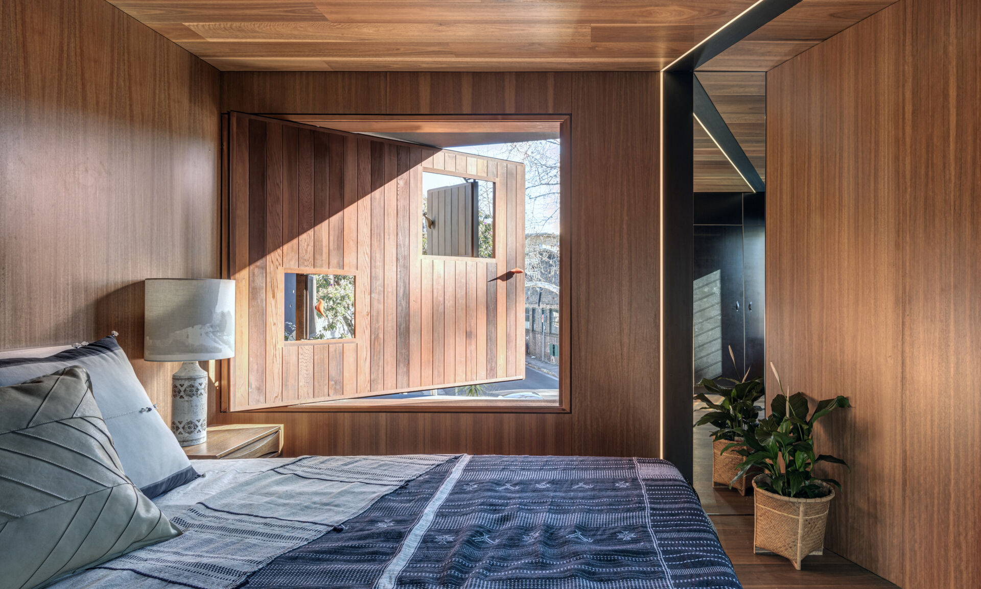 The private areas of the home, like this bedroom, feels warm and enveloping, with rich timber paneling covering every surface. Soft morning light filters through a pivoting window, offering views outside while preserving privacy.