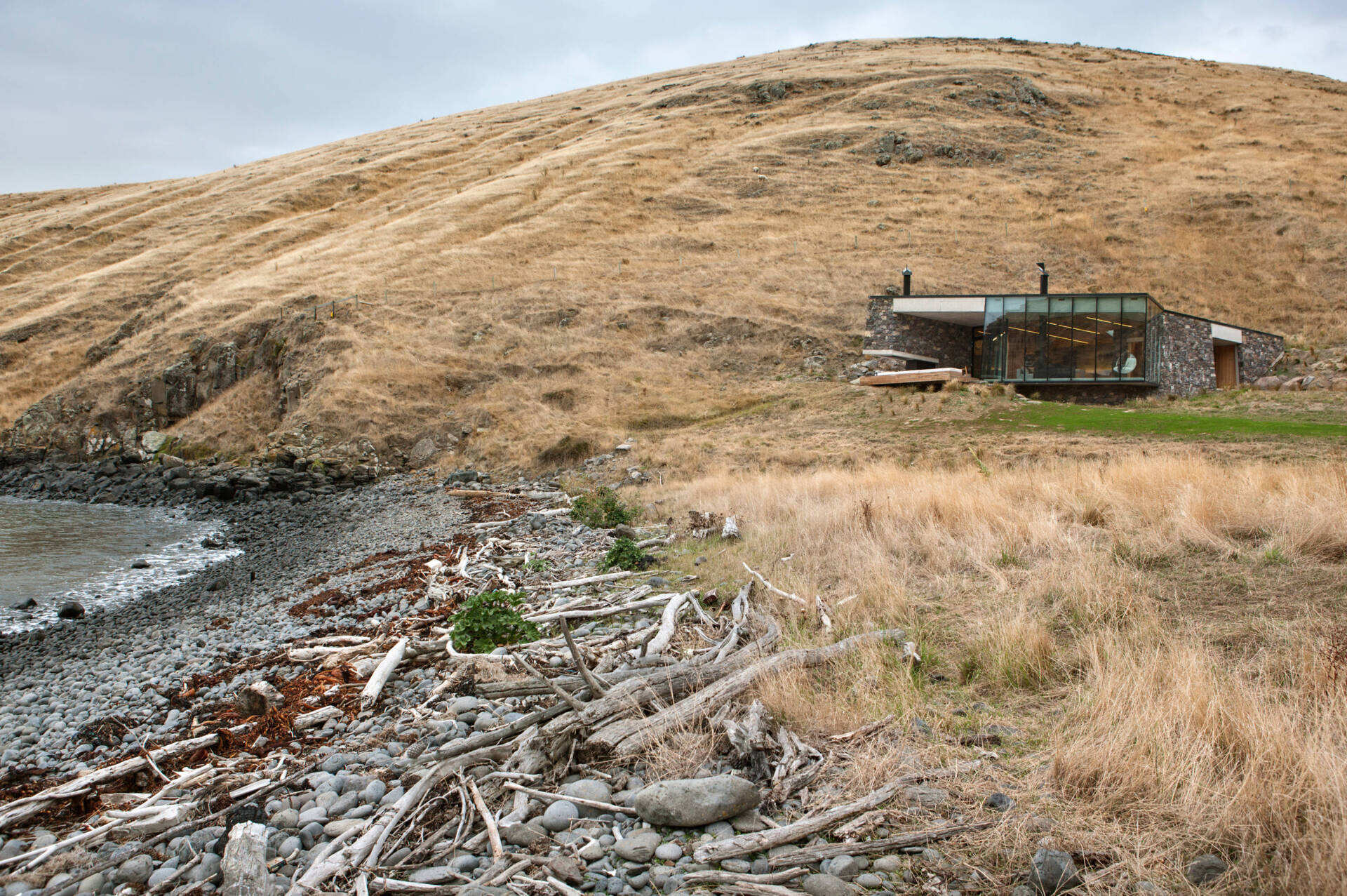 A stone cottage built using local rock, concrete, and timber to disappear into the landscape. The green roof and interlocking plan create a seamless connection with sea and sky.