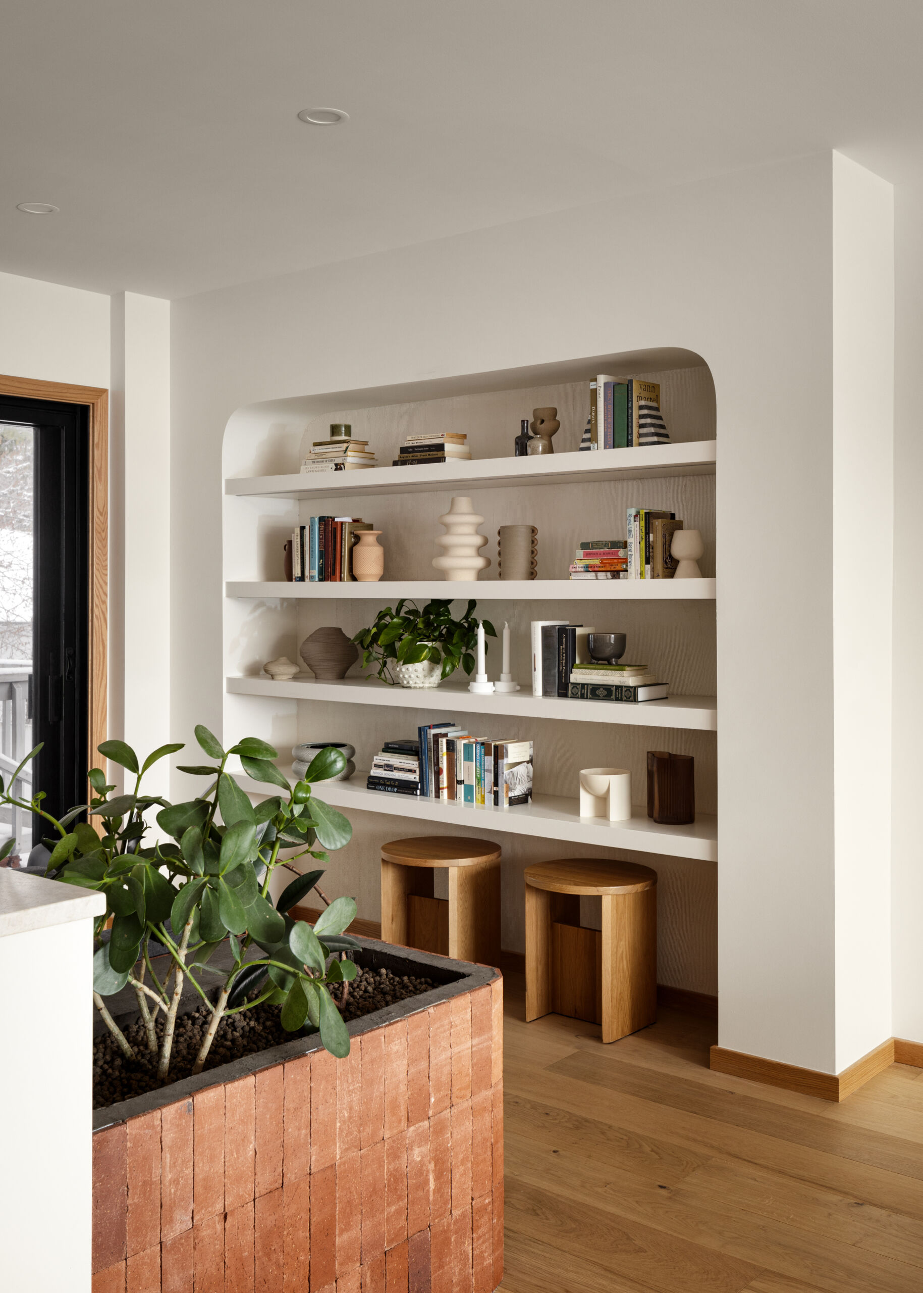 Tucked into a softly curved wall niche, the reading nook combines sculptural shelving with quiet utility. Built-in shelves hold a curated mix of books, ceramics, and plants, blending practicality with a gallery-like aesthetic.