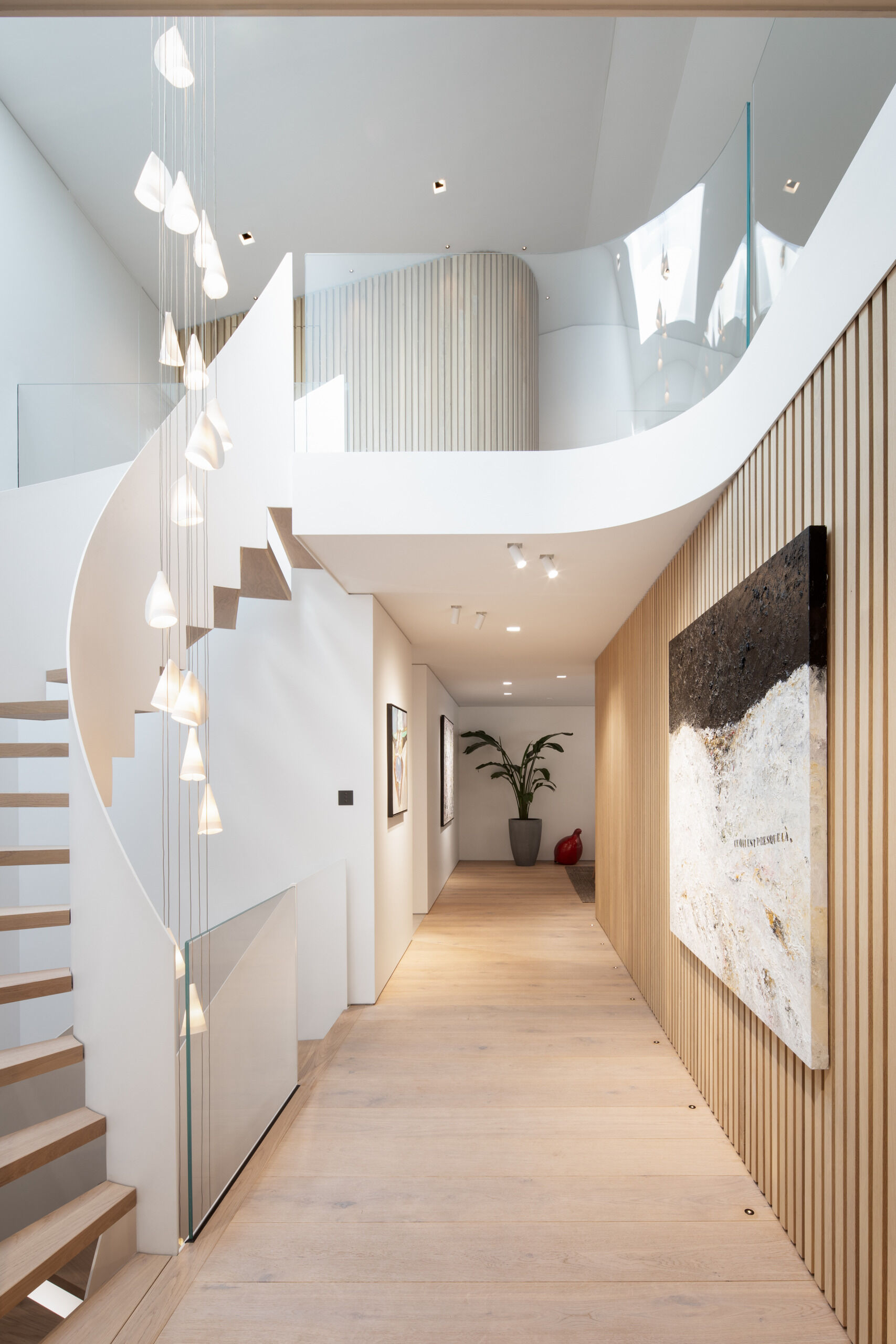 Soft curves, warm wood, and glowing light define the sculptural stairwell. Pale oak treads spiral upward beside a cascade of delicate pendants, echoing the curve with gentle rhythm, while slatted walls and smooth white surfaces add texture and calm.