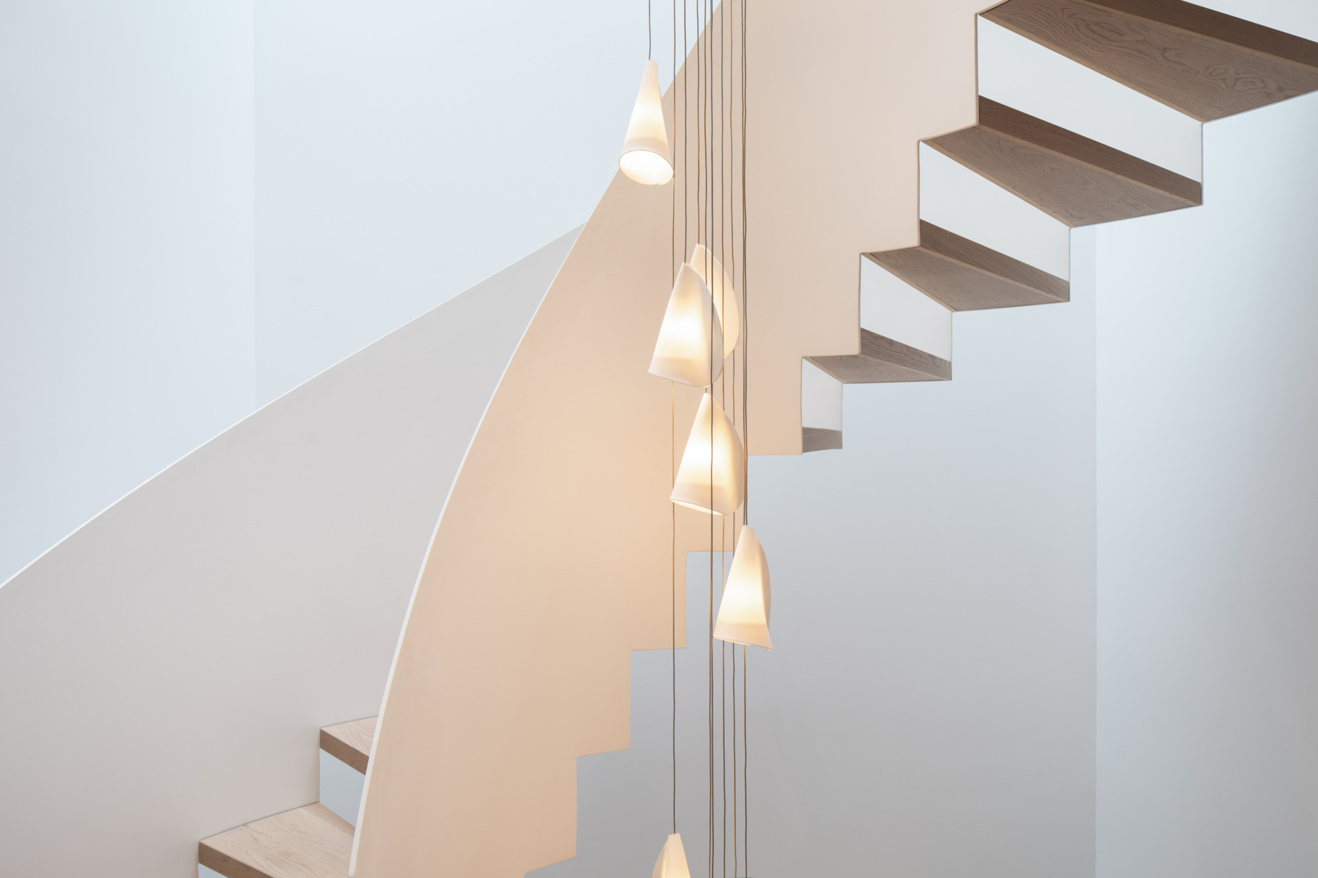 Soft curves, warm wood, and glowing light define the sculptural stairwell. Pale oak treads spiral upward beside a cascade of delicate pendants, echoing the curve with gentle rhythm, while slatted walls and smooth white surfaces add texture and calm.