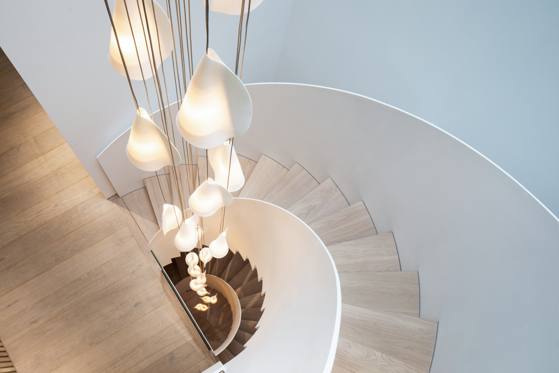 Soft curves, warm wood, and glowing light define the sculptural stairwell. Pale oak treads spiral upward beside a cascade of delicate pendants, echoing the curve with gentle rhythm, while slatted walls and smooth white surfaces add texture and calm.