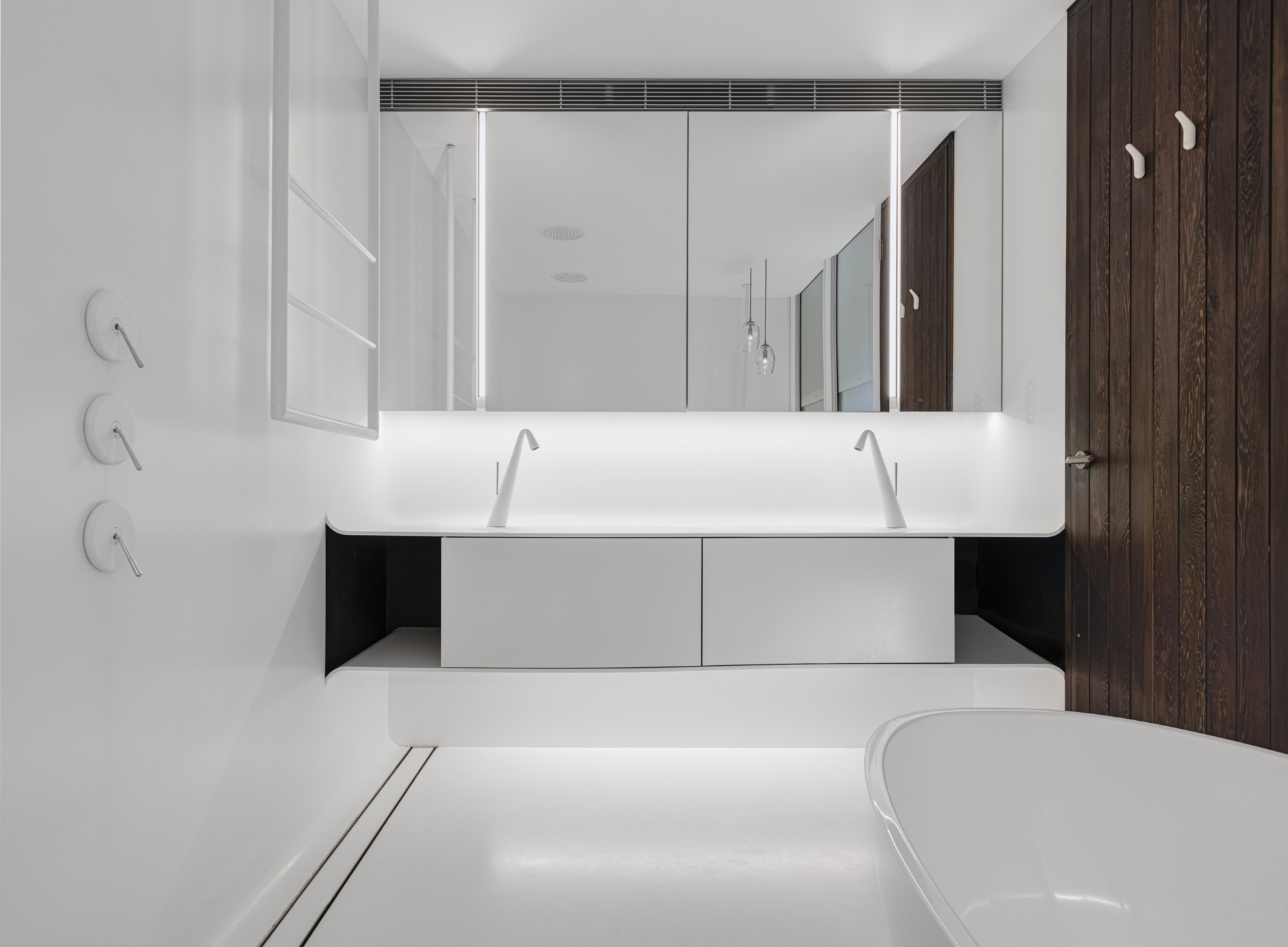 Crisp white finishes and seamless cabinetry give this bathroom a pared-back, contemporary feel. A floating double vanity and sculptural tapware reinforce the clean lines, while concealed lighting adds a soft, ambient glow. A timber door introduces warmth, offering a quiet contrast to the otherwise minimal palette.