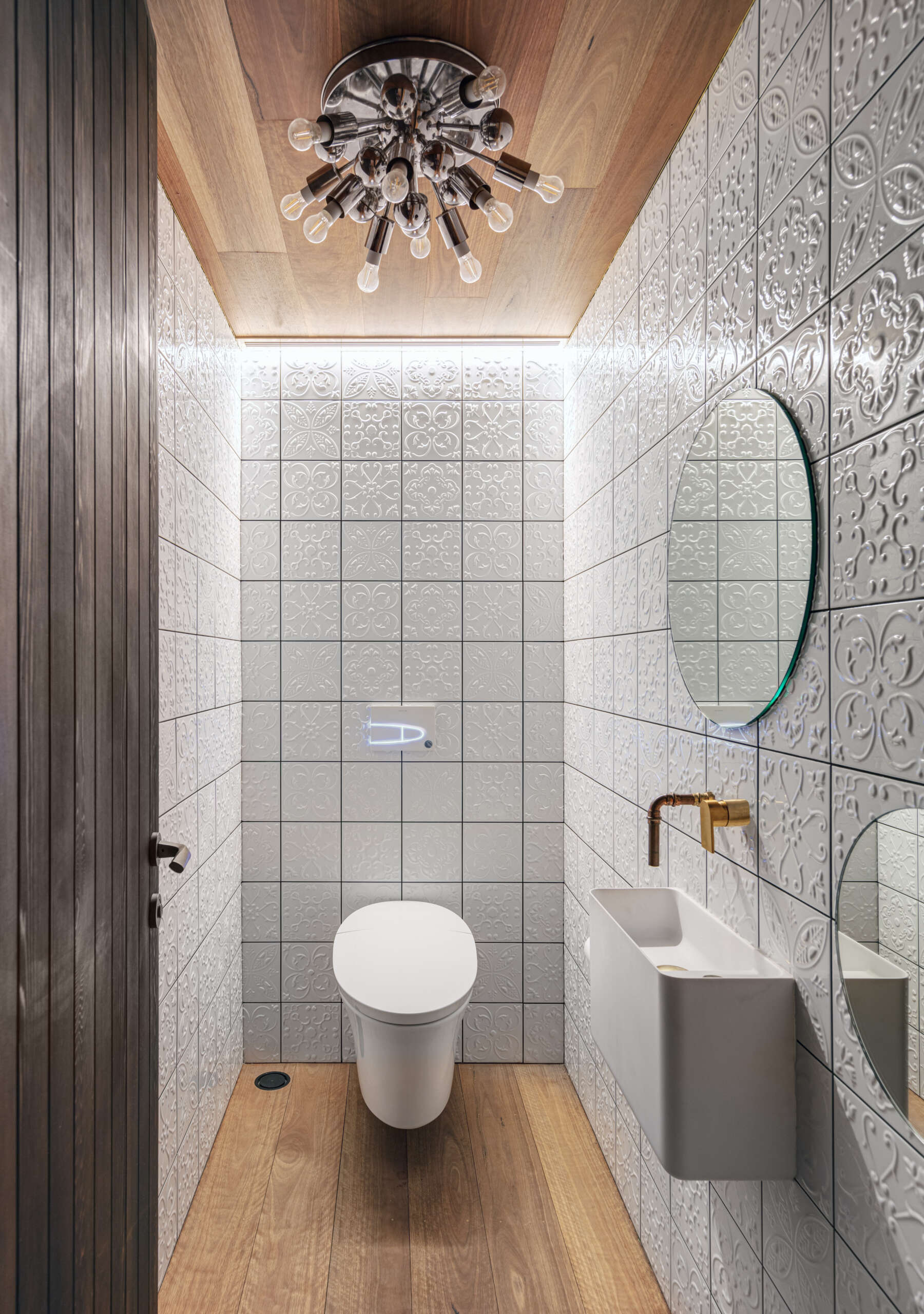 This powder room pairs subtle texture with refined detailing. Embossed white tiles wrap the walls, while timber flooring and ceiling bring warmth and contrast. A slim brass tap and minimalist basin keep the layout clean, and a sculptural light overhead adds a quiet sense of elegance to the compact space.