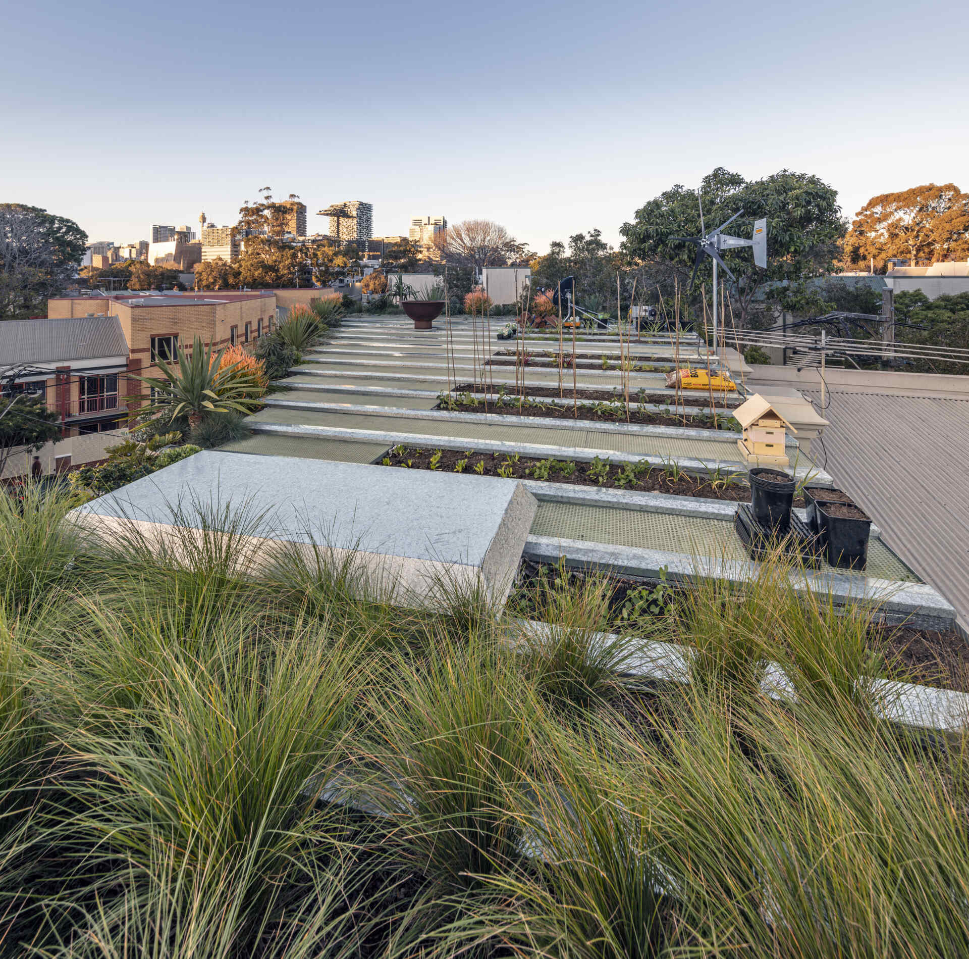 The top of the home is dedicated to a stepped garden offers a tranquil green refuge amid the city, with organized planting beds for herbs and vegetables. Grasses move gently beside clean stone paths, while raised beds and small wind turbines underscore a sustainable approach. At sunset, a sculptural fire bowl anchors the terrace, inviting quiet moments framed by hardy plants and expansive skyline views.