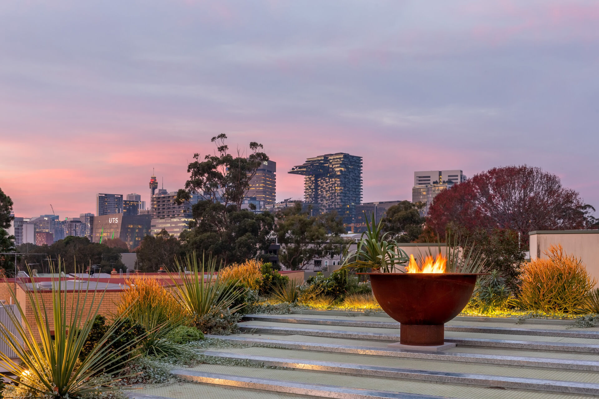 The top of the home is dedicated to a stepped garden offers a tranquil green refuge amid the city, with organized planting beds for herbs and vegetables. Grasses move gently beside clean stone paths, while raised beds and small wind turbines underscore a sustainable approach. At sunset, a sculptural fire bowl anchors the terrace, inviting quiet moments framed by hardy plants and expansive skyline views.