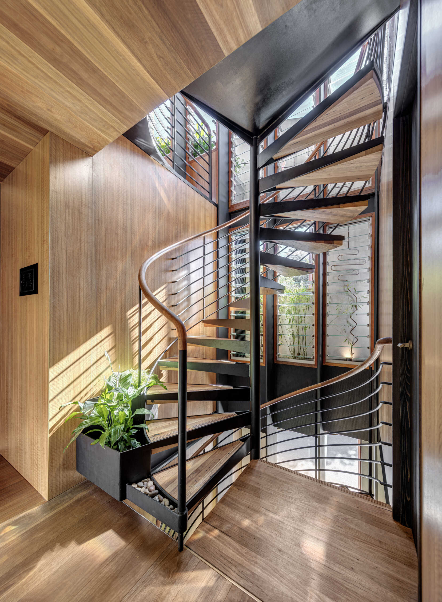 A spiral staircase brings sculptural beauty to a sunlit corner of the home, with its graceful wood steps and sleek black railing wrapping upward in an elegant curve. Floor-to-ceiling windows flood the space with light, highlighting the warm tones of the wood and the lush greenery planted at the stair’s base.