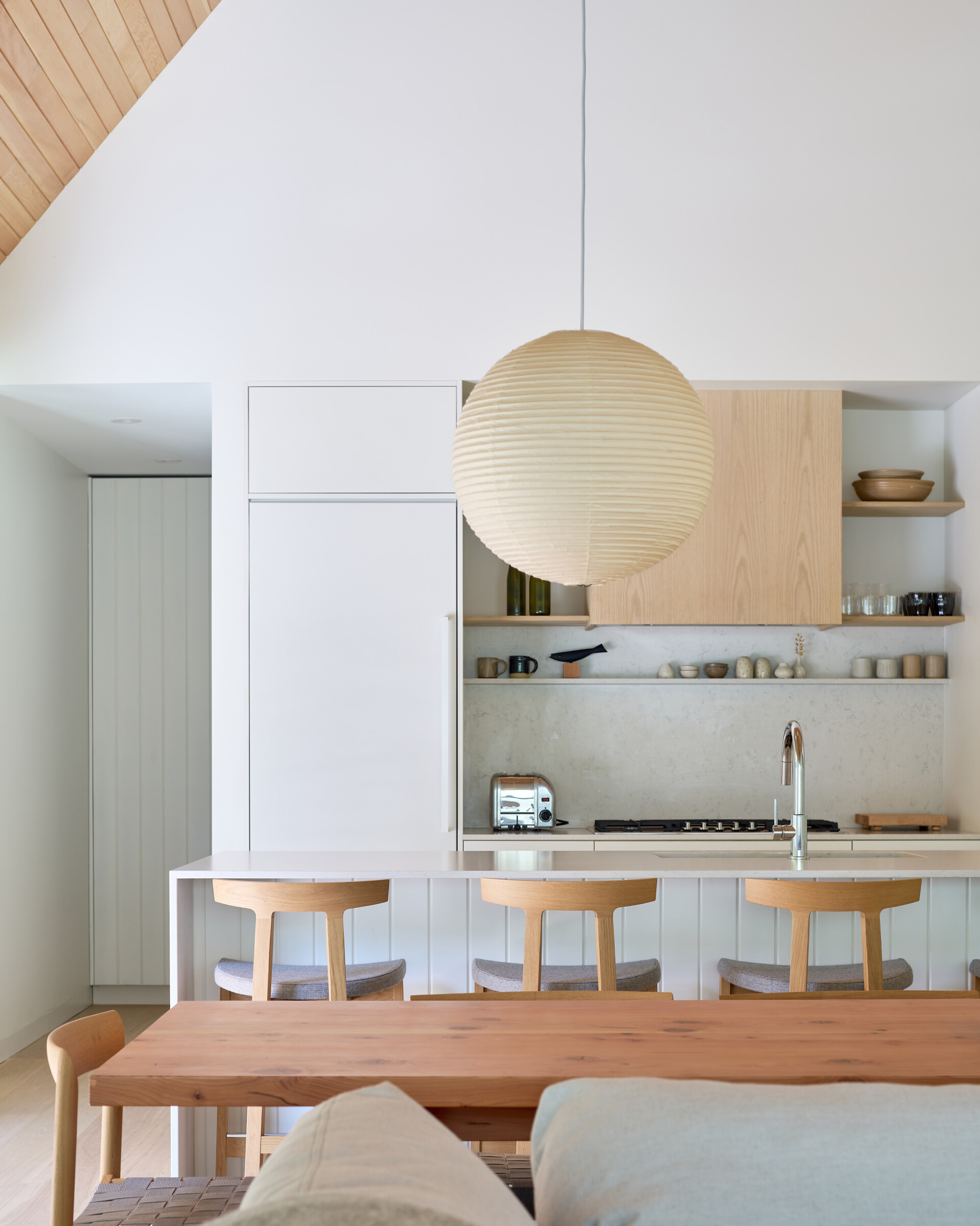 This dining and kitchen area is anchored by a long wood table paired with simple chairs, creating a warm and inviting place for meals. The kitchen features a clean palette of light wood cabinetry, open shelving, and a pale backsplash, offering both function and calm simplicity. A central island with seating adds versatility, serving as a spot for casual dining or gathering while cooking.