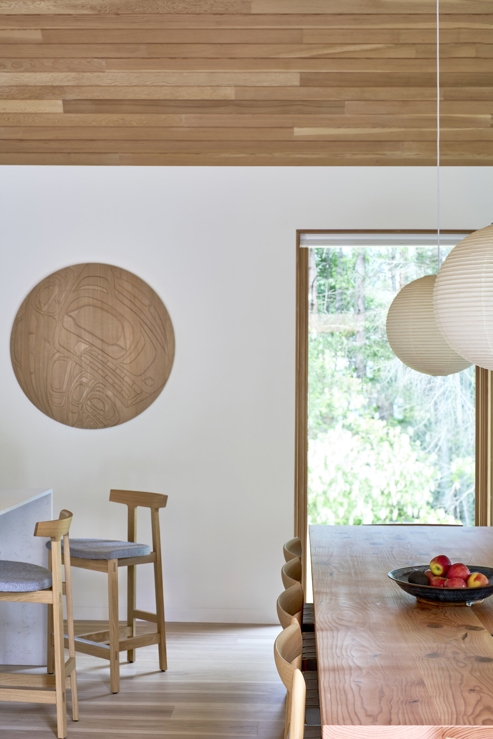 This dining and kitchen area is anchored by a long wood table paired with simple chairs, creating a warm and inviting place for meals. The kitchen features a clean palette of light wood cabinetry, open shelving, and a pale backsplash, offering both function and calm simplicity. A central island with seating adds versatility, serving as a spot for casual dining or gathering while cooking.