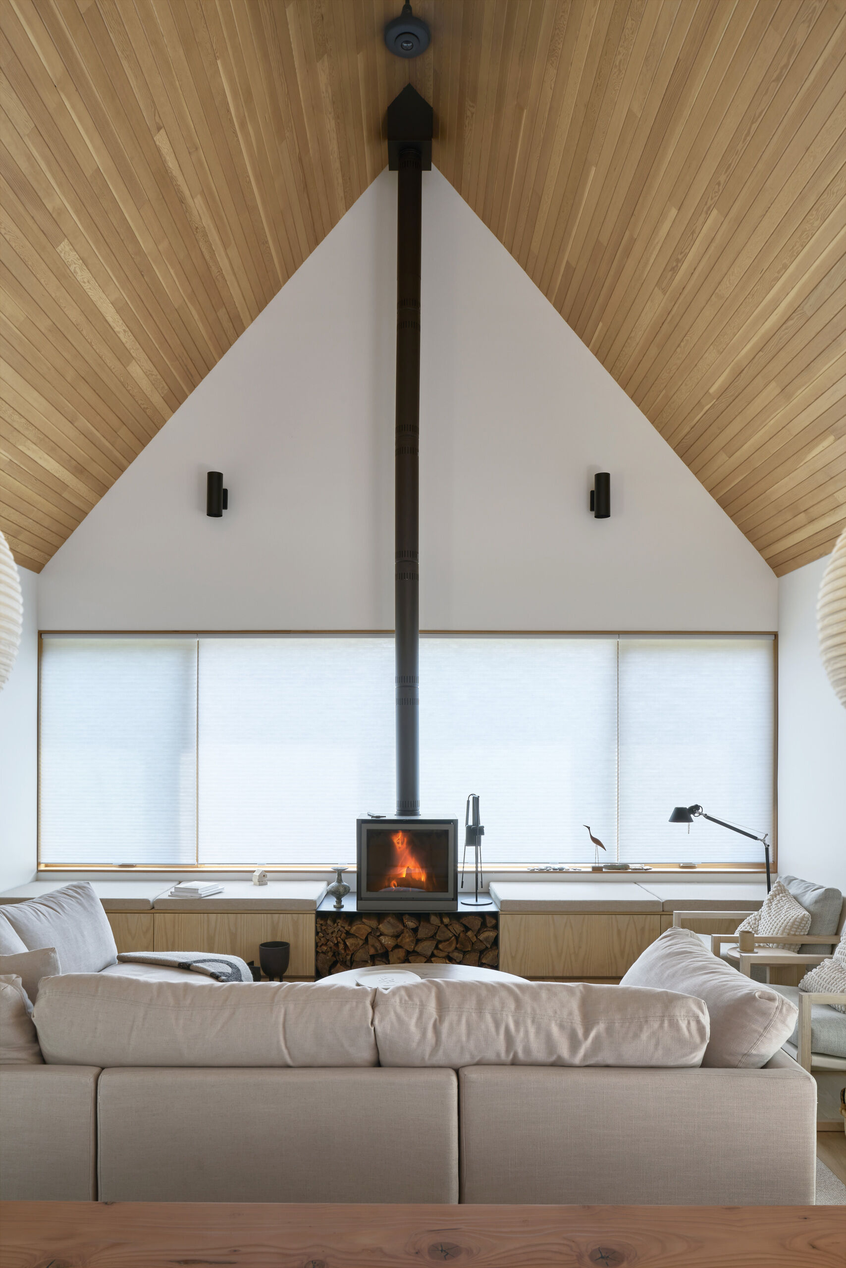 Inside, the living room is designed as a flexible gathering space, where windows frame views of the surrounding forest. A wood-burning fireplace anchors the room, bringing both warmth and atmosphere. Built-in benches run along the windows, doubling as seating and storage, and keeping the space streamlined.