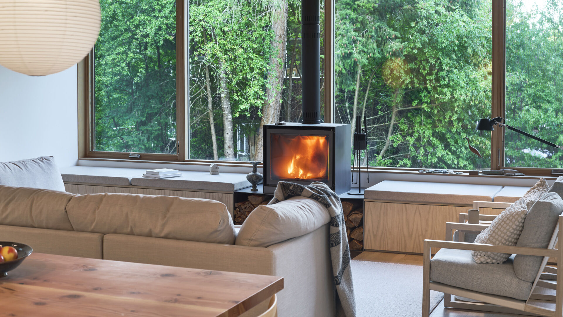 Inside, the living room is designed as a flexible gathering space, where windows frame views of the surrounding forest. A wood-burning fireplace anchors the room, bringing both warmth and atmosphere. Built-in benches run along the windows, doubling as seating and storage, and keeping the space streamlined.