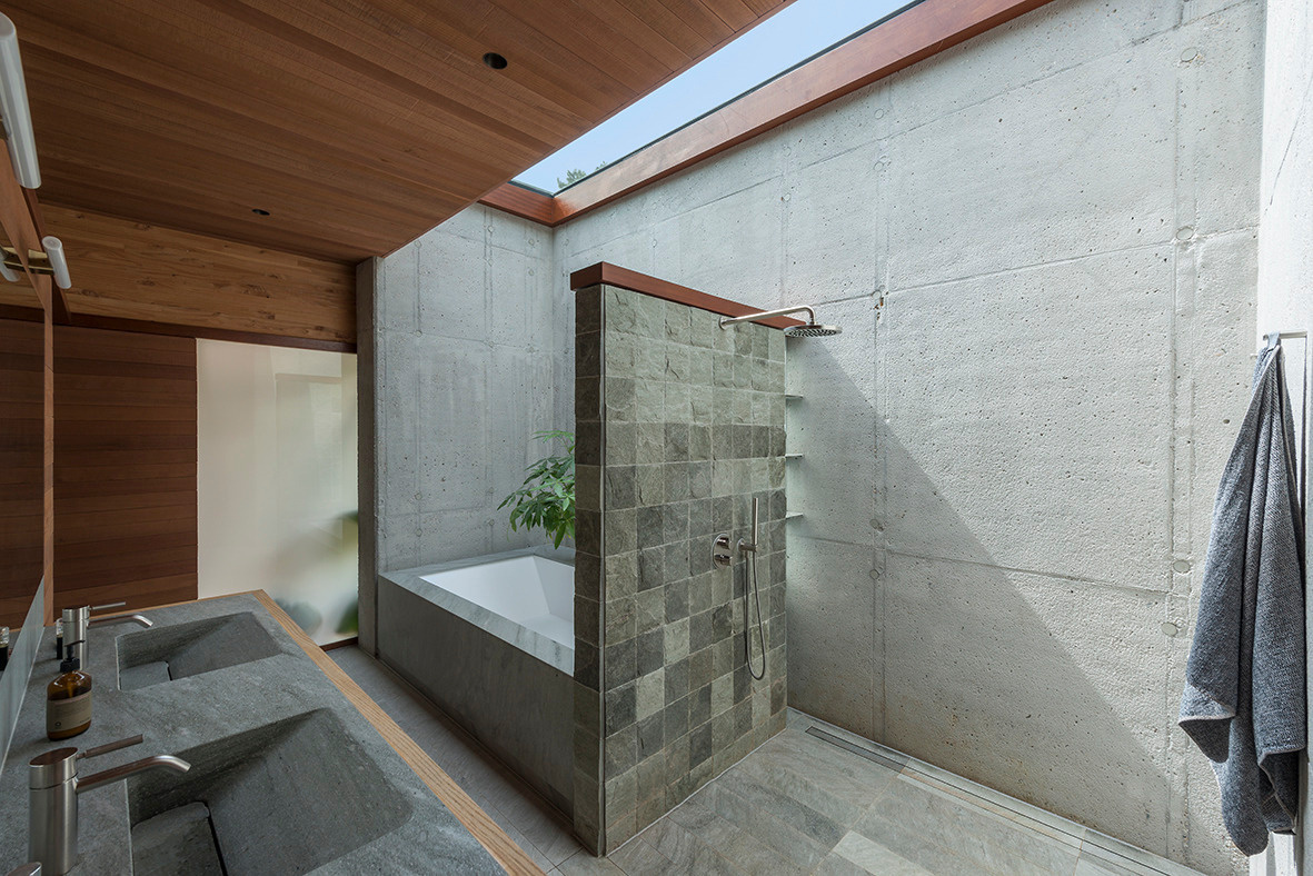 This bathroom combines raw materials and refined details for a clean, modern look. Concrete walls and stone tile surfaces create a natural, grounded feel, while a large skylight floods the space with daylight. The layout features a deep soaking tub tucked behind a tiled partition, paired with an open walk-in shower for a seamless, spa-like flow. Dual sinks in carved stone add a sculptural touch, and the overall design reflects the home’s balance of functionality, warmth, and understated luxury.