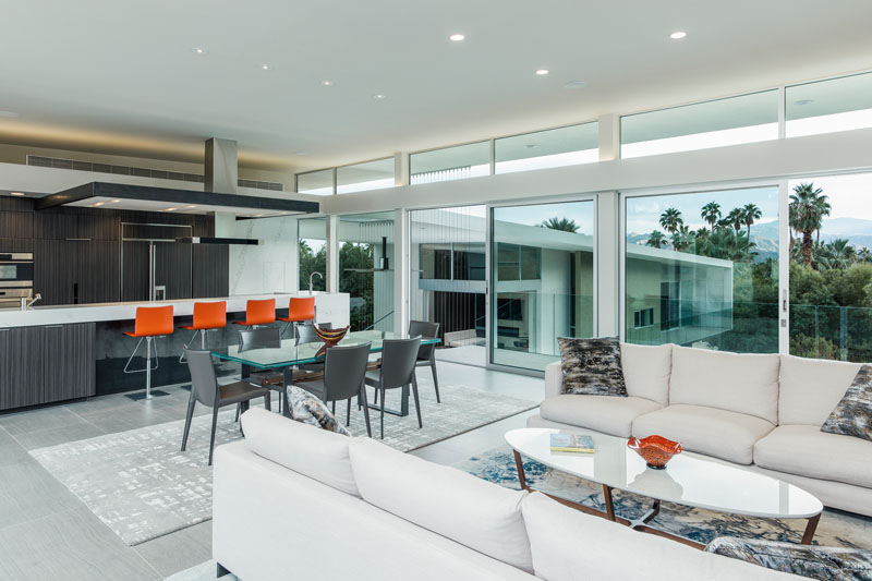 The living room, dining area, and kitchen share one continuous space. Floor-to-ceiling glass walls maximize views of the surrounding desert and mountains, making the landscape a constant presence in daily life.