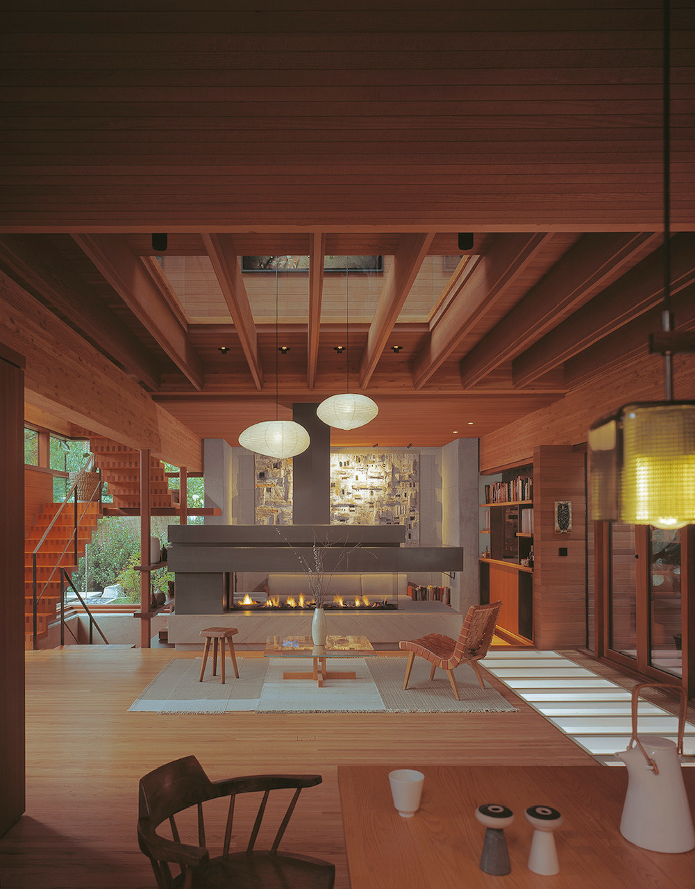 This living room revolves around a double-sided fireplace, providing both visual drama and cozy warmth. The space feels open yet intimate, thanks to the balance of large windows, exposed structural elements, and careful furniture placement.