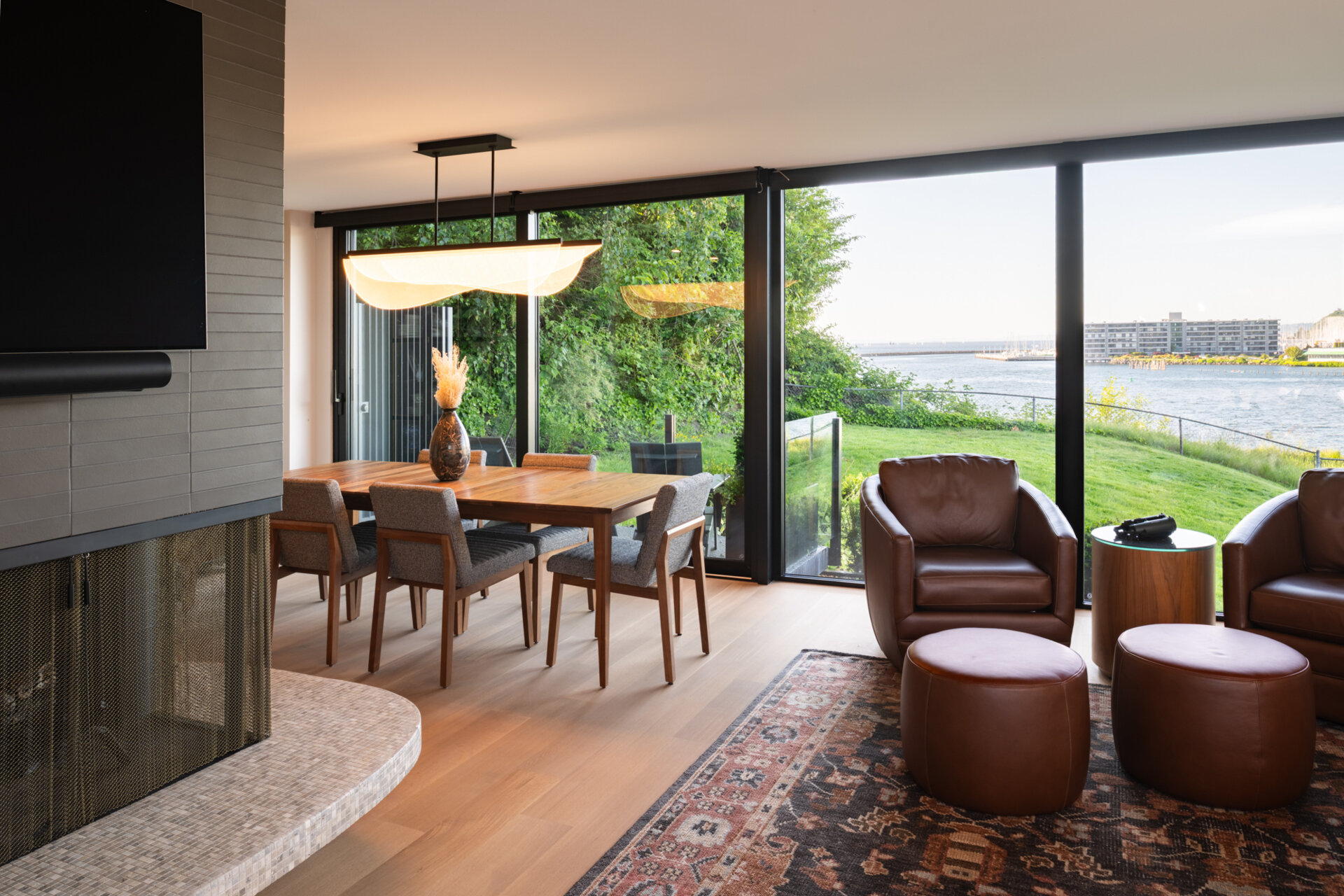 The open living and dining area flows directly from the kitchen, framed by glass walls that draw in the surrounding landscape. The original fireplace anchors the room, while the updated finishes provide a fresh backdrop for gatherings. The effect is seamless continuity between past and present.