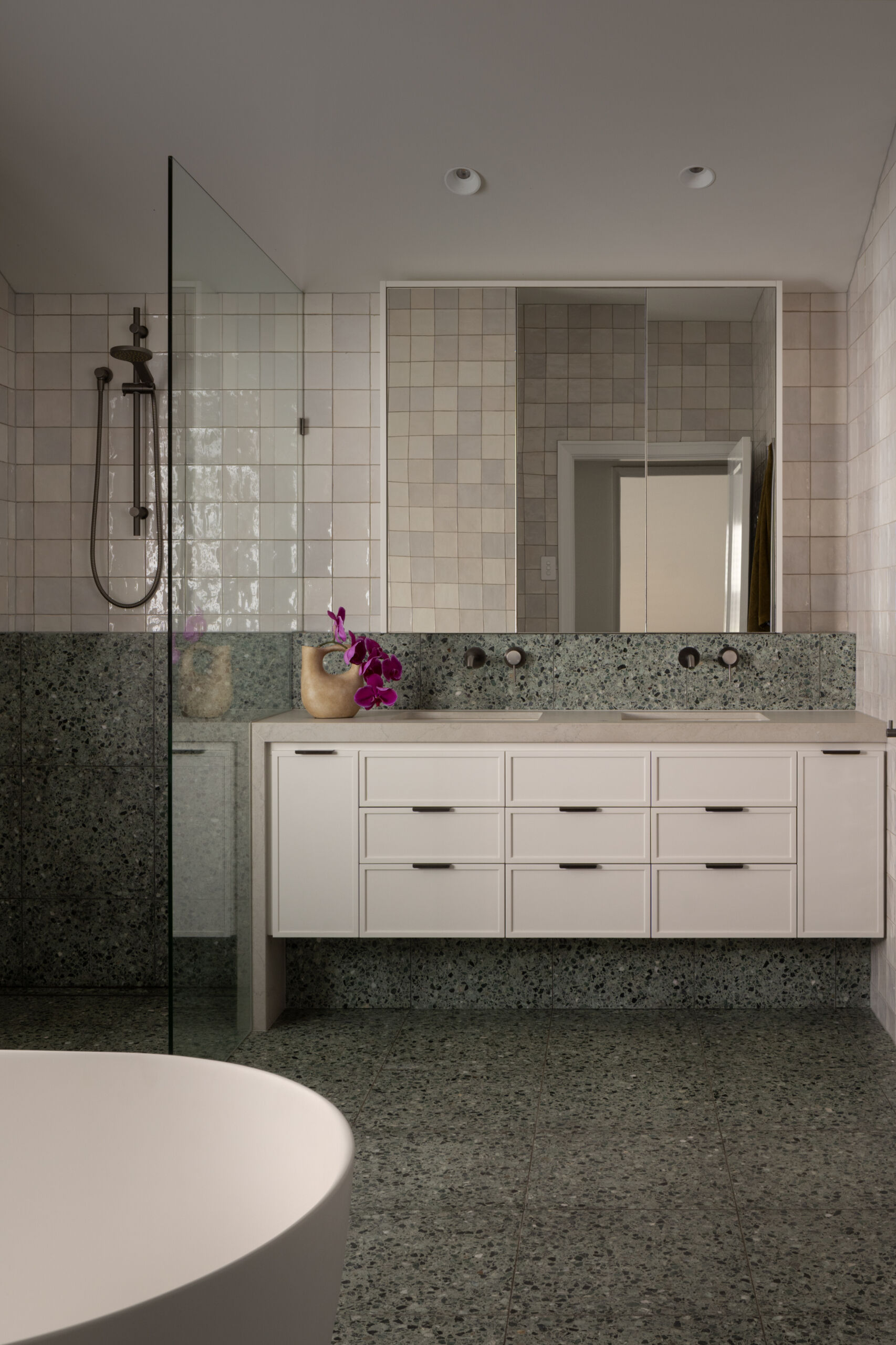 Soft neutral tones and layered textures give this bathroom a calm, inviting feel. Pale square tiles line the walls in a subtle patchwork of cream and beige, while speckled green terrazzo underfoot introduces an earthy, grounded note. A white vanity keeps the space light, paired with slim black hardware for definition.