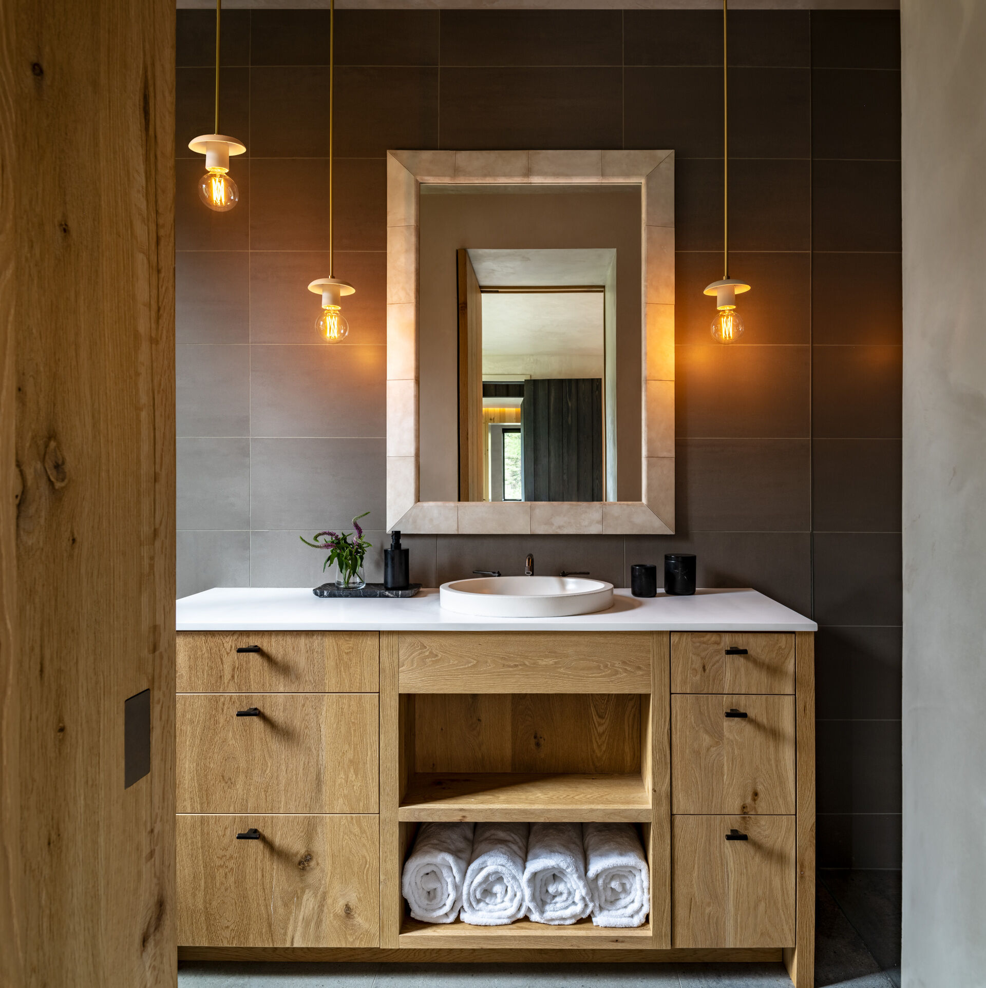 This bathroom blends warmth and modern simplicity. The natural wood vanity feels inviting with its clean grain and open shelving. A crisp white countertop and round sink keep the look fresh, while soft gray wall tiles add depth without overpowering the space, and hanging pendant lights cast a golden glow beside the stone-framed mirror.